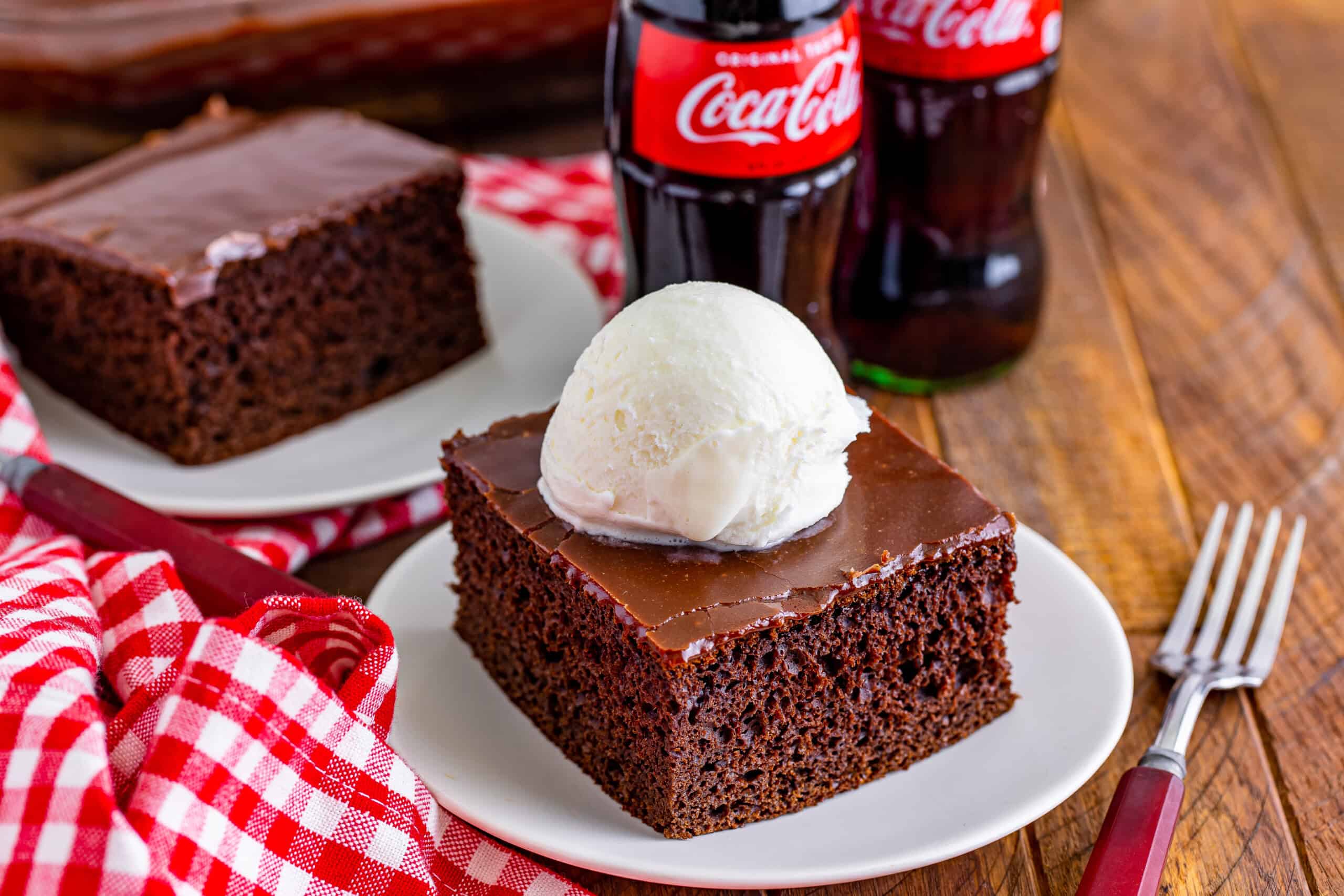 The image features a slice of chocolate cake accompanied by a scoop of vanilla ice cream on a plate. Beside the plate, there is a red checkered napkin and two bottles of Coca-Cola.