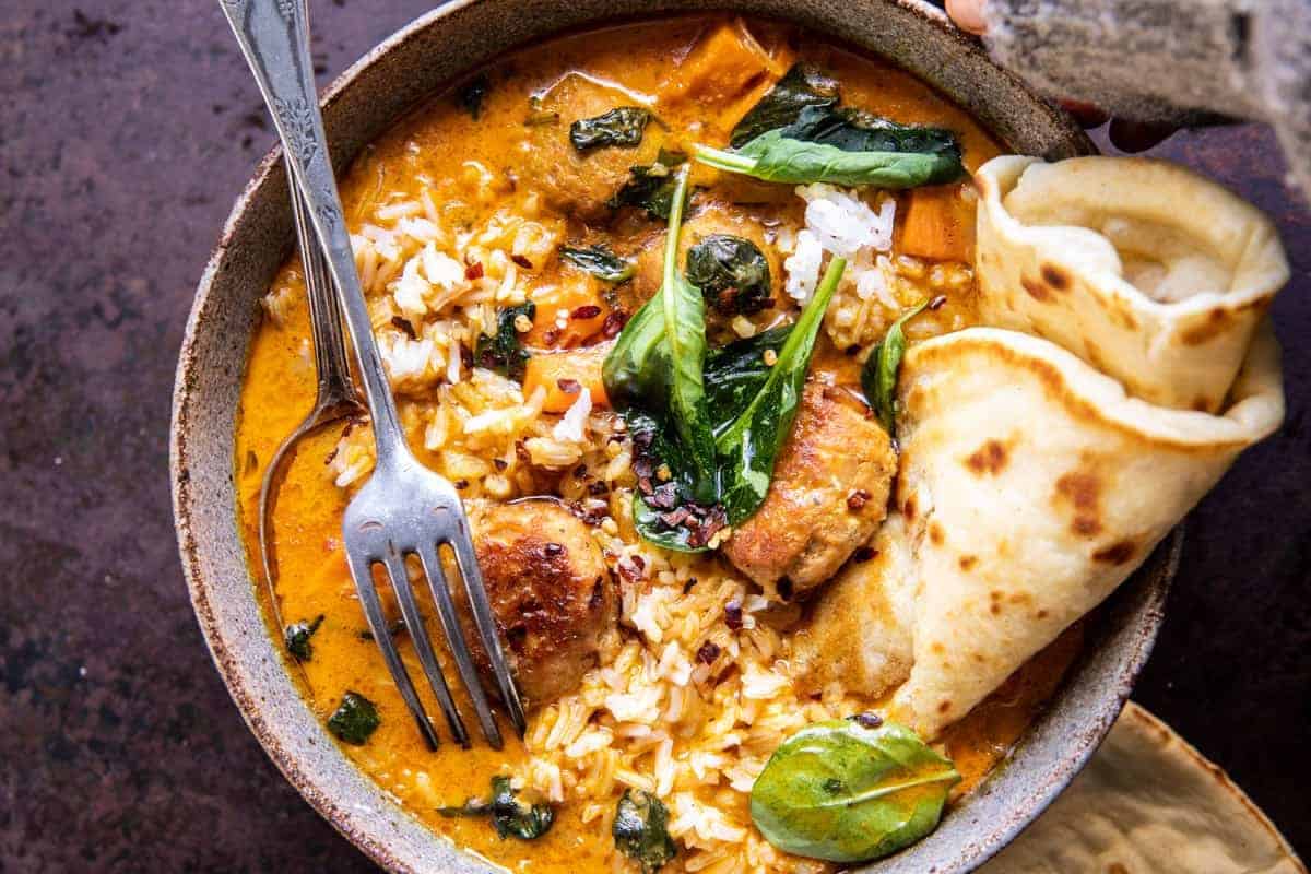 A bowl of curry with chicken, rice, and leafy greens, garnished with red chili flakes. A folded piece of naan bread is on the side, and a fork rests in the bowl. The rich, colorful dish sits on a dark, textured surface.