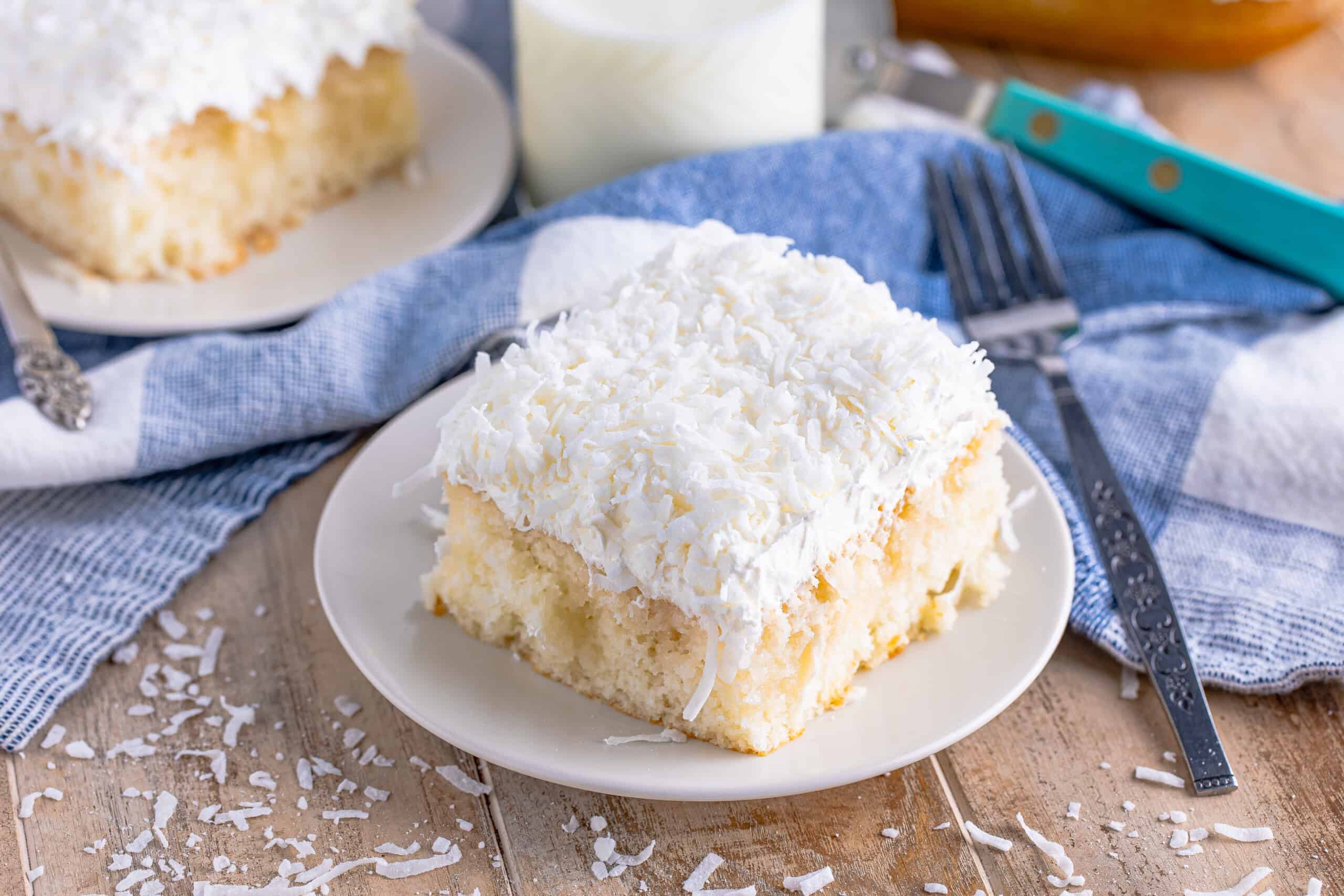 A slice of coconut cake on a white plate, topped with shredded coconut. A fork and a knife rest nearby on a blue and white cloth.