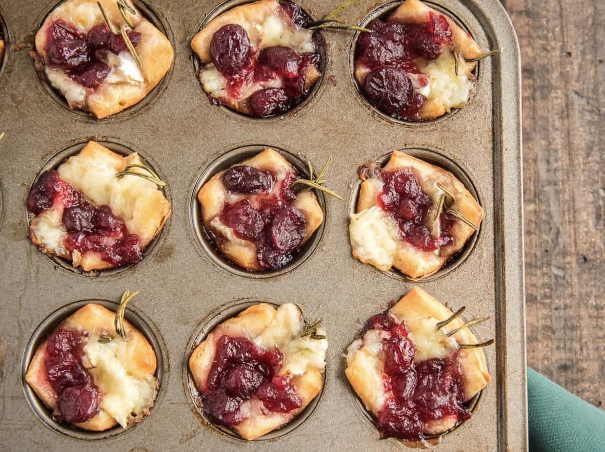 A muffin tray with baked pastries topped with melted cheese, cherry toppings, and garnished with rosemary sprigs.