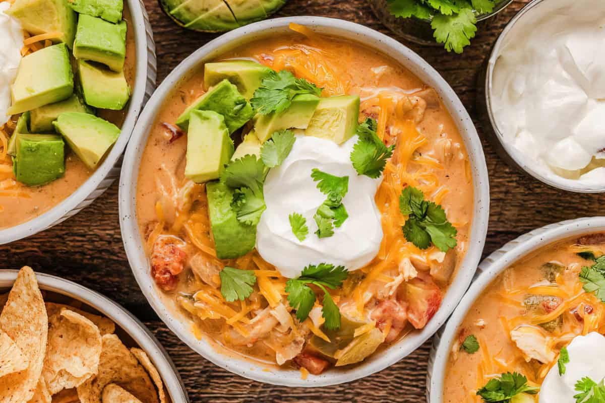 A bowl of creamy soup topped with avocado slices, sour cream, cilantro leaves, and shredded cheese. It is accompanied by tortilla chips and extra toppings around the bowl.
