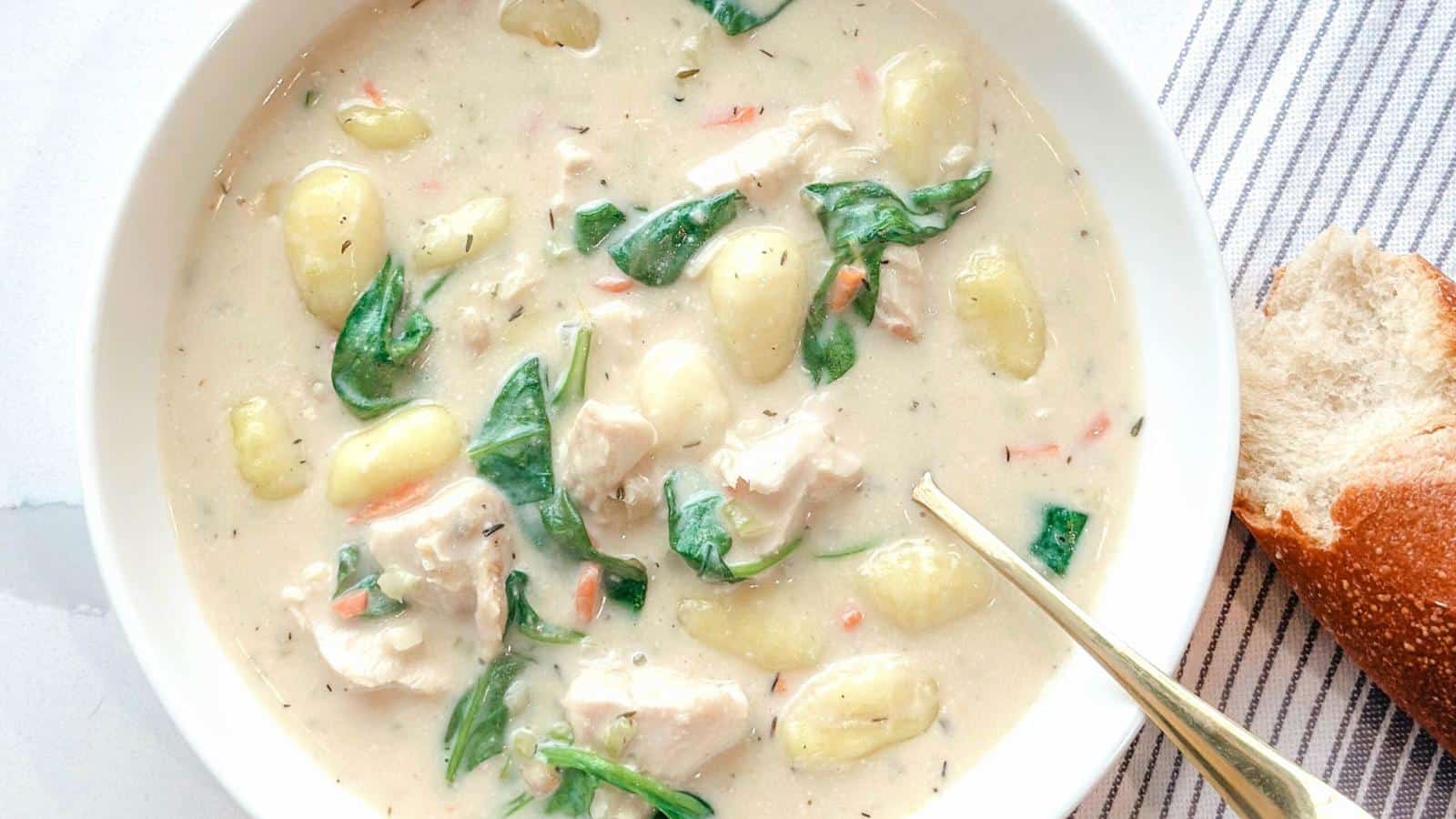 The image shows a bowl of creamy soup containing gnocchi, chicken pieces, spinach, and carrots, with a piece of bread placed beside it.