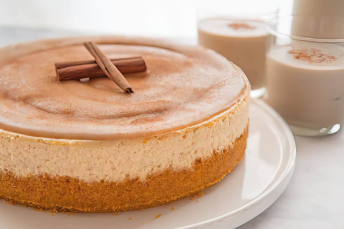 A delicious cheesecake topped with cinnamon sticks on a white plate. Two glasses of creamy beverage with a sprinkle of cinnamon are in the background, creating a cozy and inviting atmosphere.