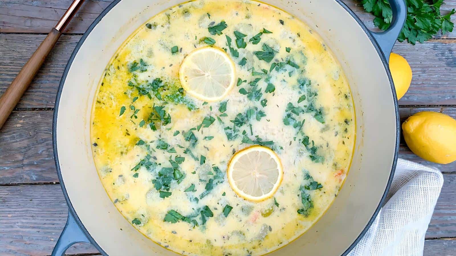 A pot of creamy soup garnished with lemon slices and chopped herbs. The soup is in a round pot placed on a wooden surface, with a few lemons and a kitchen towel nearby.