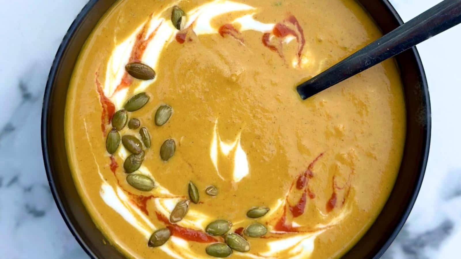 A bowl of pumpkin soup with swirls of cream, pumpkin seeds, a drizzle of red sauce, and a spoon inside.