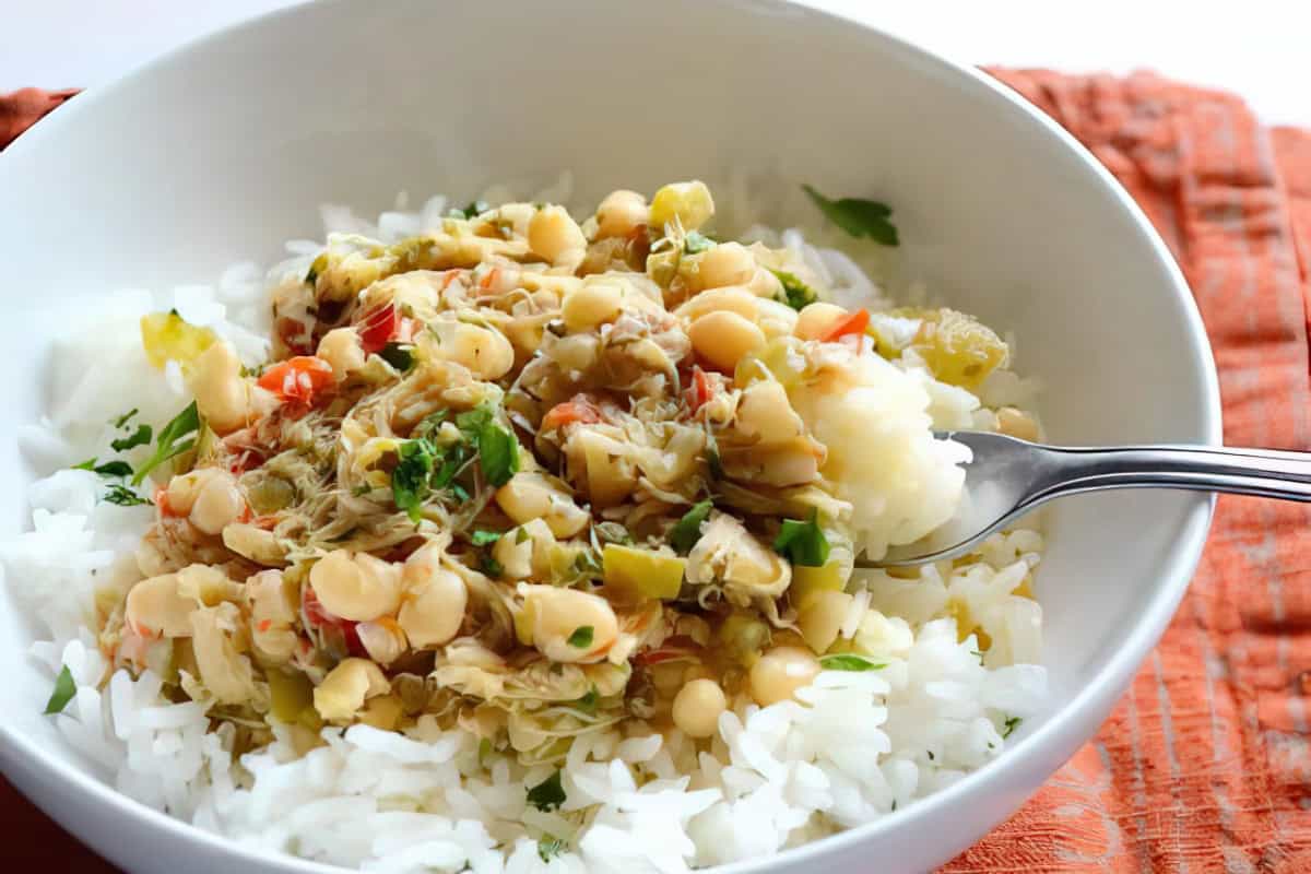 A bowl of white rice topped with a colorful mixture of beans, shredded vegetables, and herbs. A fork is placed in the bowl, ready to take a bite. The dish is set on an orange cloth.