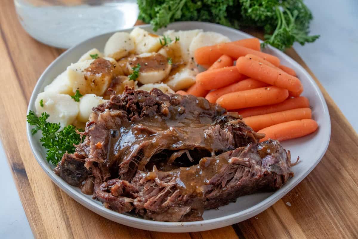 A plate featuring roast beef with gravy, accompanied by baby carrots and sliced potatoes, garnished with fresh parsley.