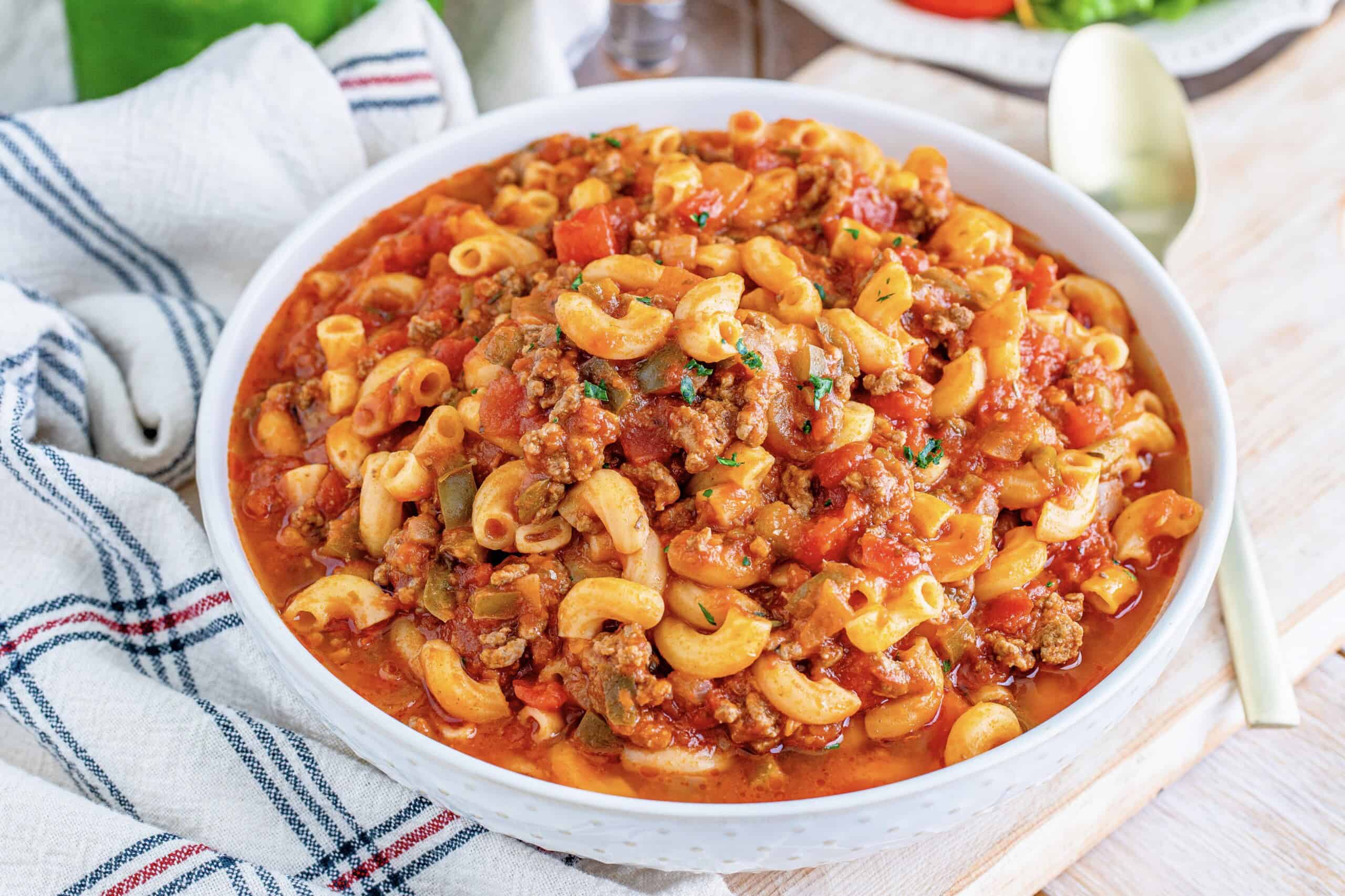 A bowl of macaroni pasta with ground beef and tomato sauce, a napkin, and a spoon on a table.