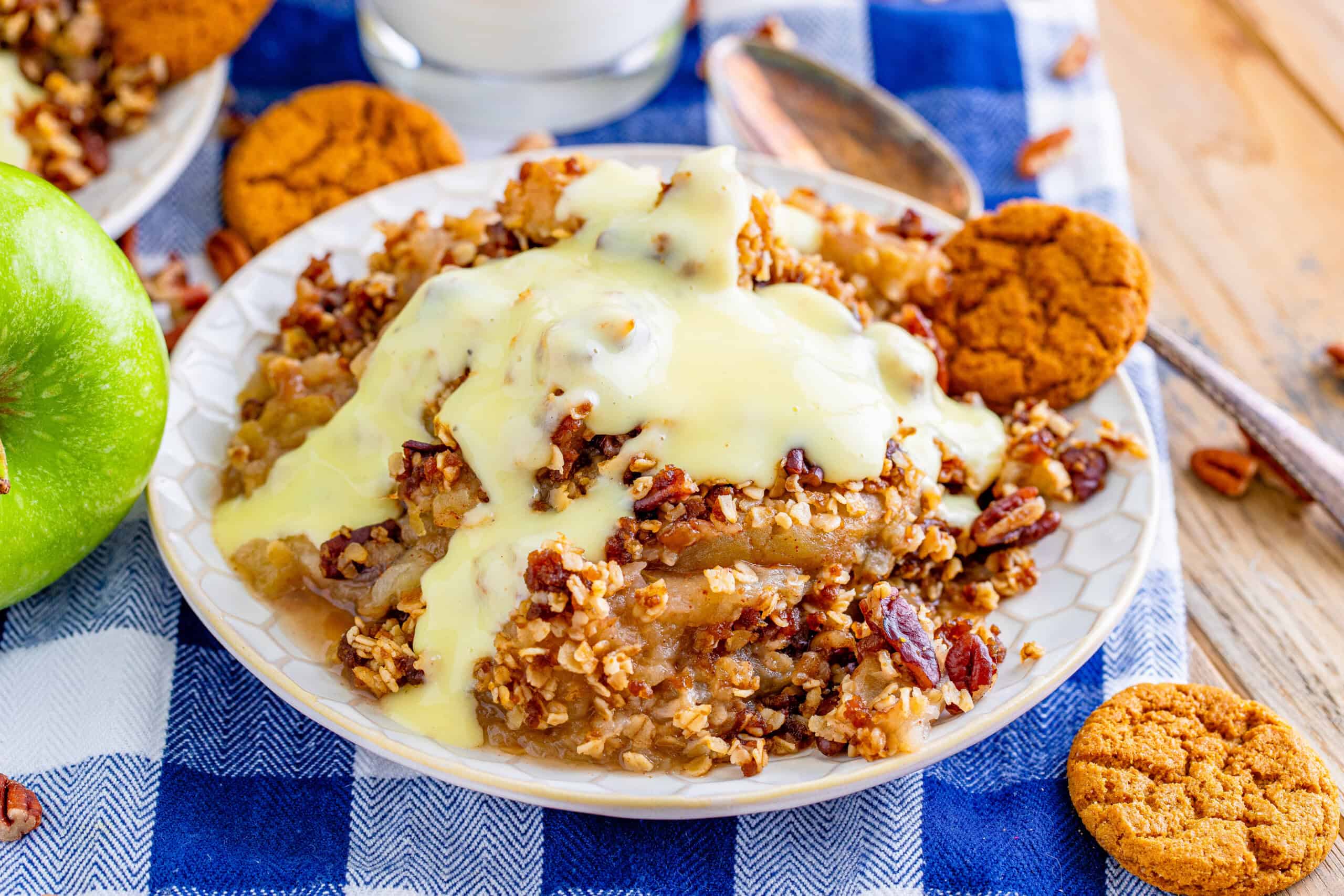 The image features a plate of apple crisp topped with vanilla sauce and garnished with pecans, surrounded by cookies on a blue and white checkered cloth. In the background, there is a green apple and a glass of milk.