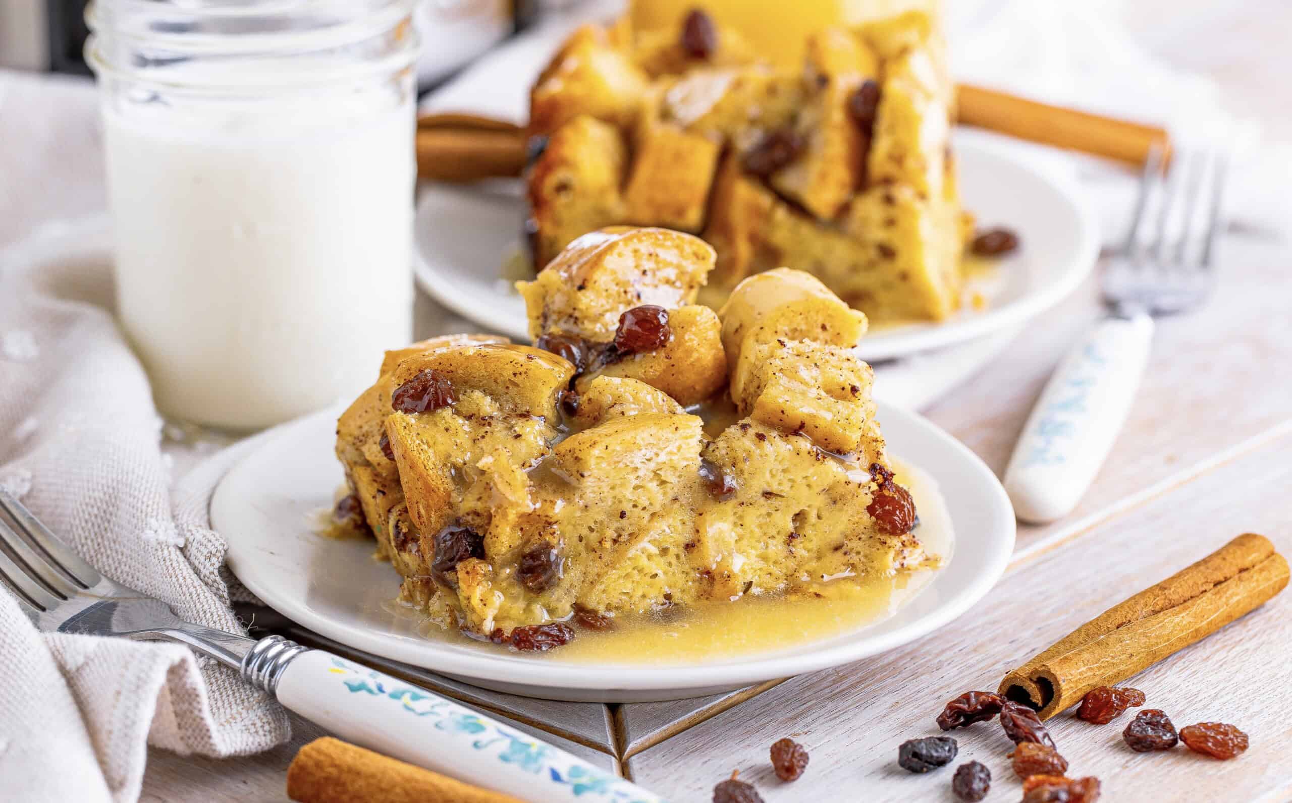 The image features two plates of bread pudding with raisins and sauce, accompanied by forks, a cinnamon stick, and a jar of milk on the table.