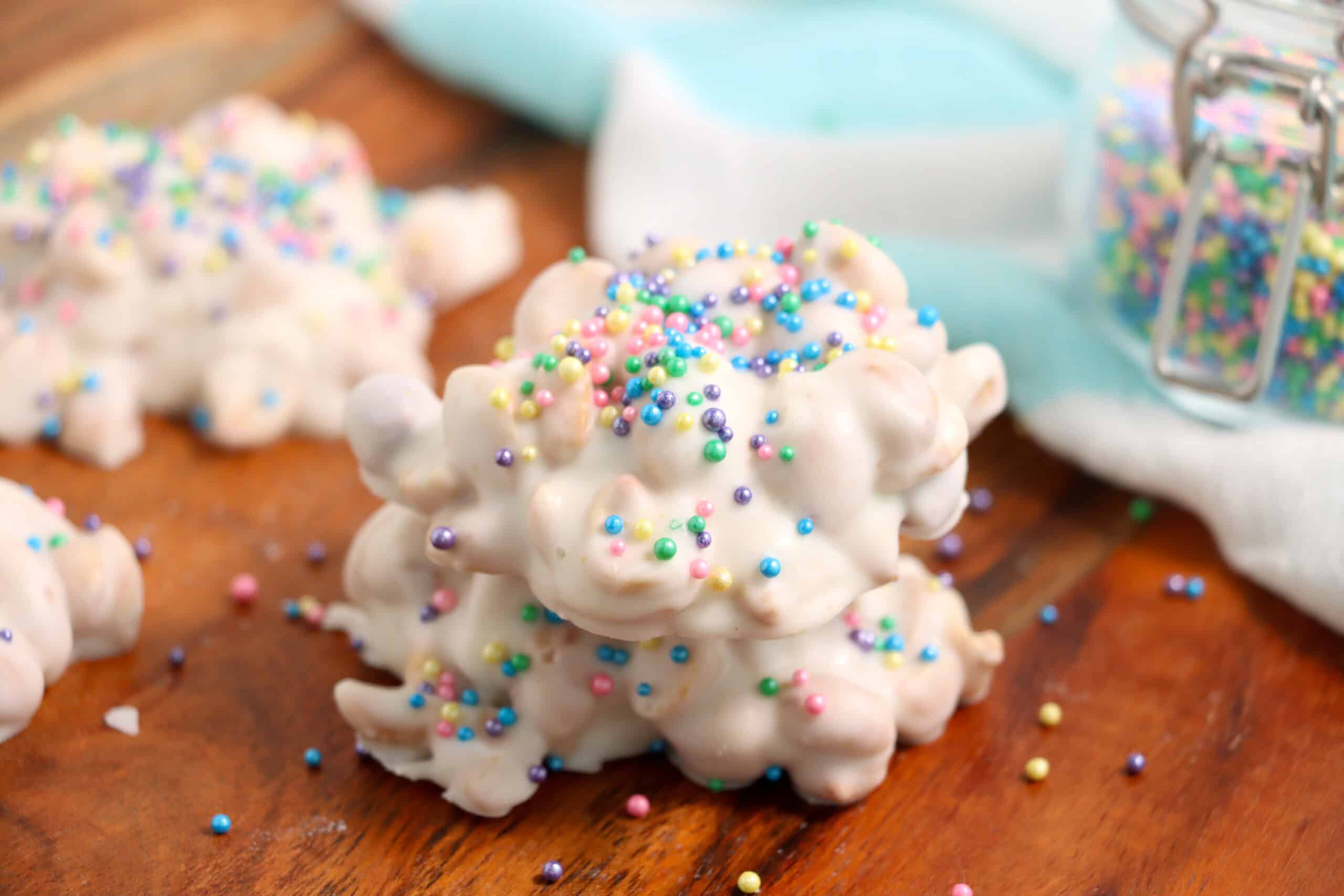 A pile of clusters coated in white chocolate and topped with colorful round sprinkles, sitting on a wooden surface.