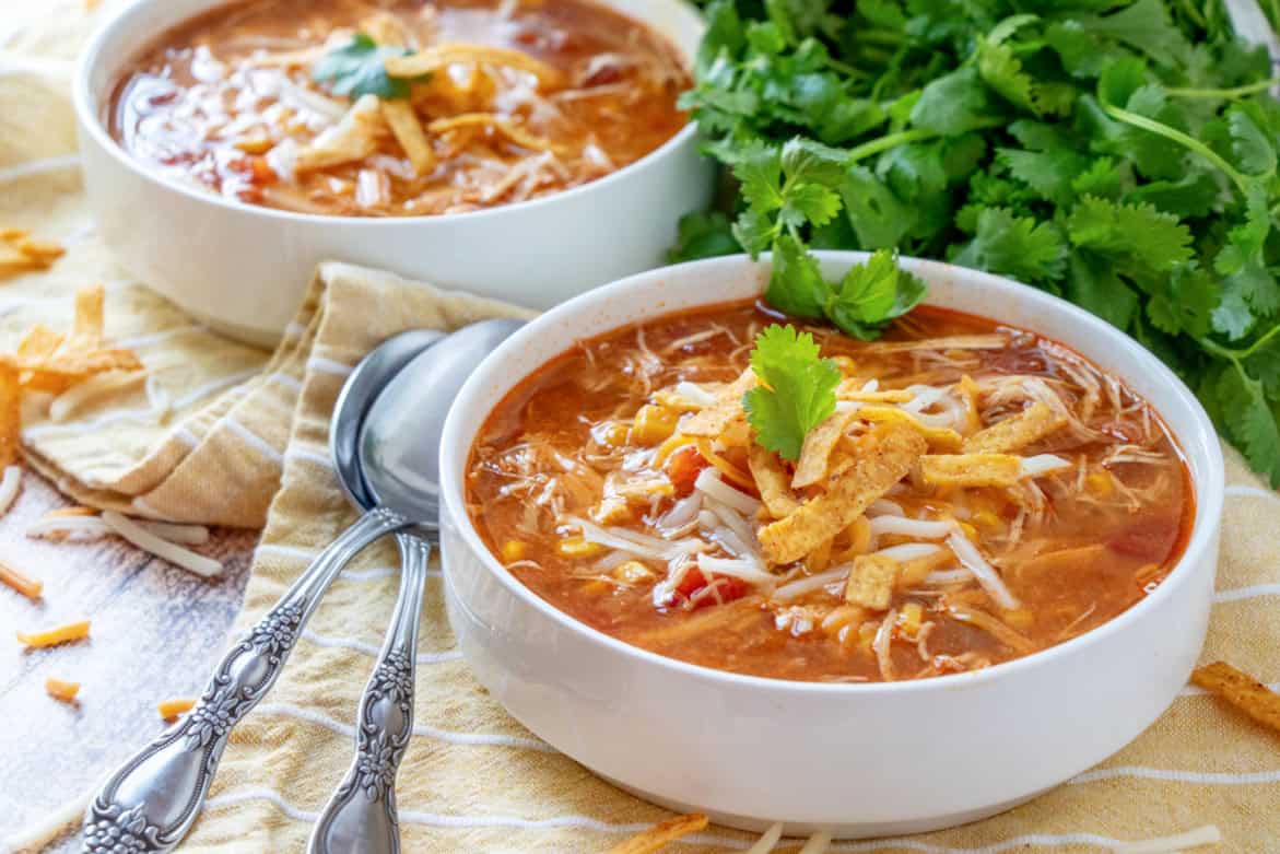 The image shows two bowls of tortilla soup topped with cheese, tortilla strips, and cilantro, placed on a striped cloth. Adjacent to the bowls are spoons and a bunch of fresh cilantro.