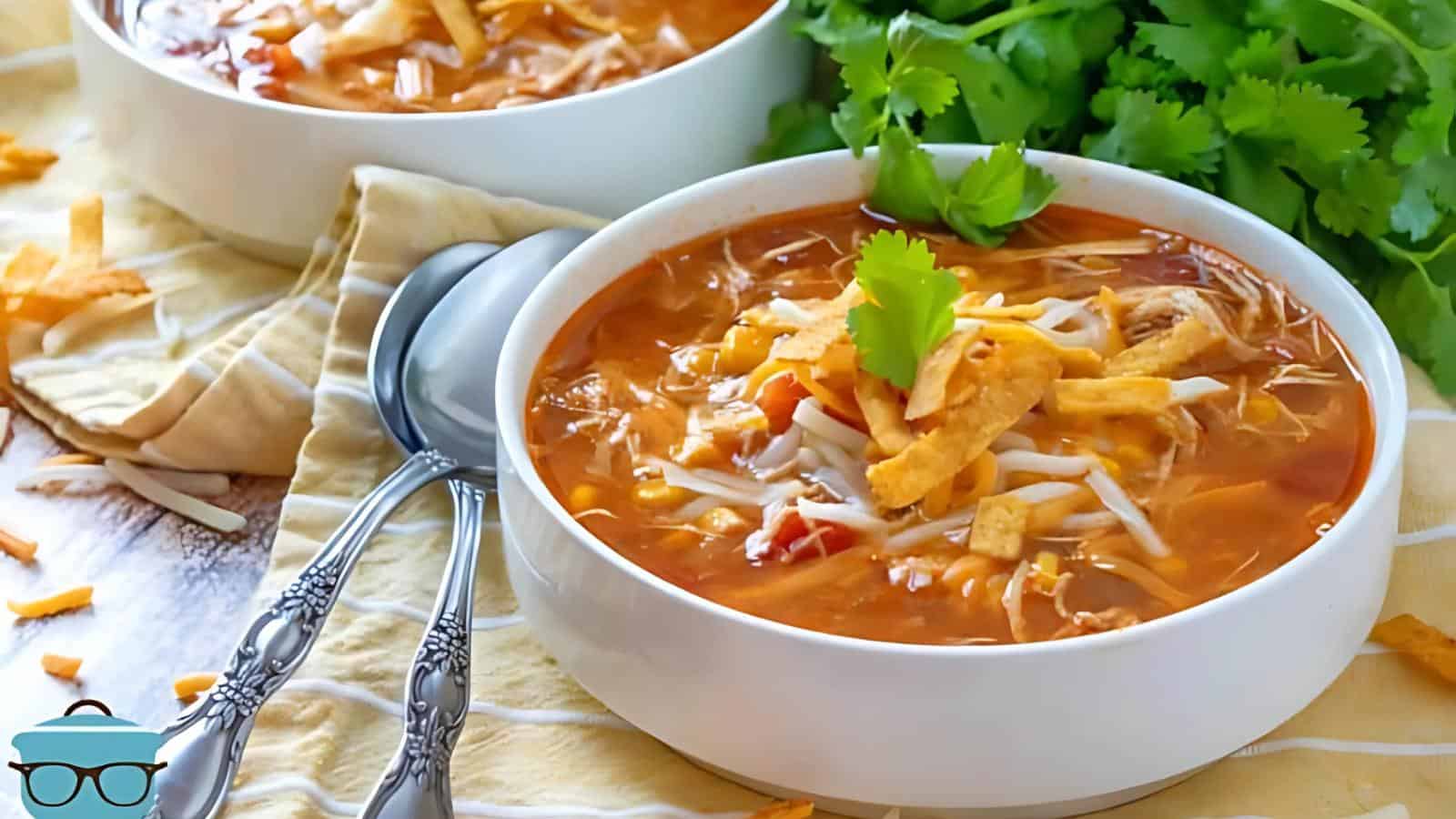 Two bowls of tortilla soup garnished with cheese and cilantro, placed on a cloth next to silver spoons and cilantro.