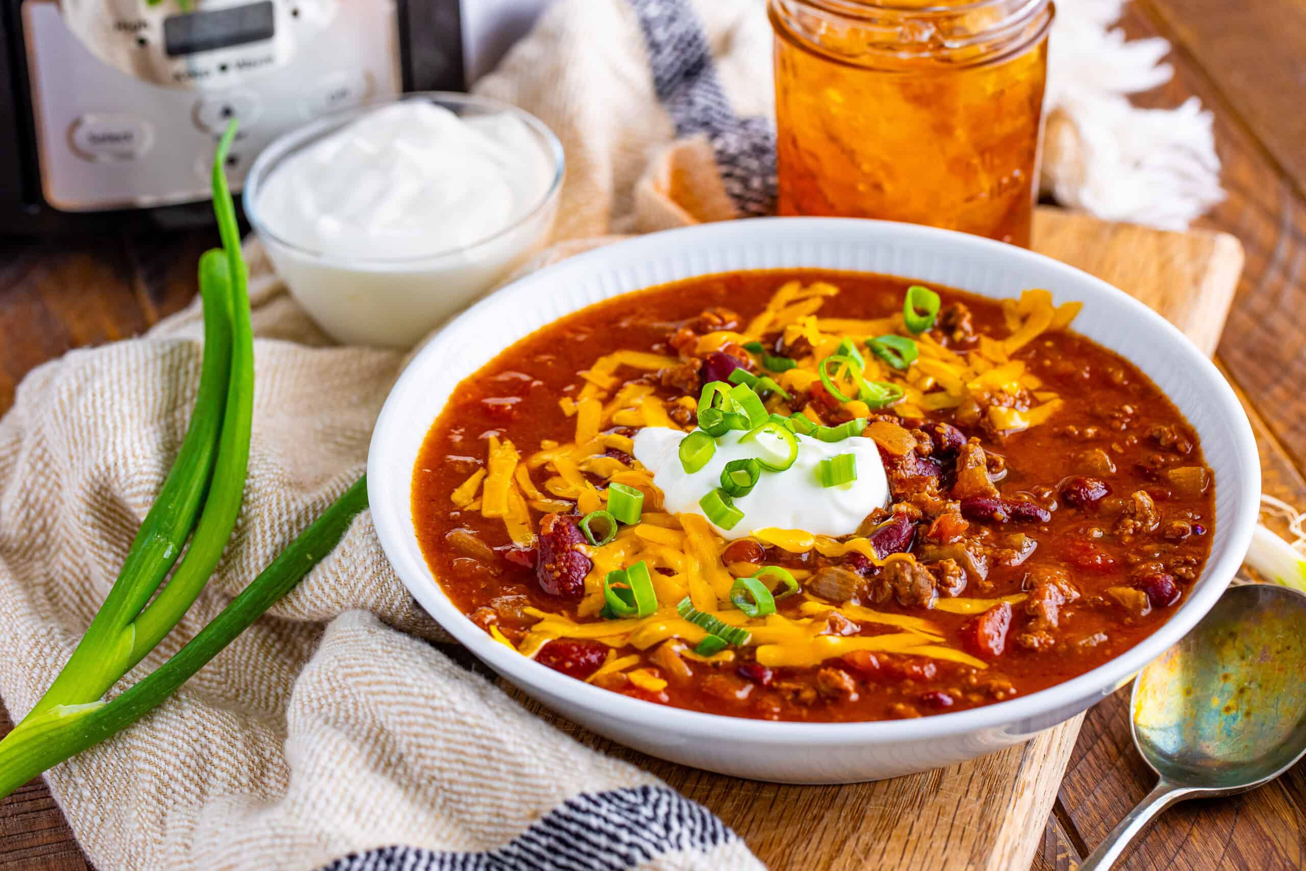 The image depicts a bowl of chili garnished with cheese, sour cream, and green onions. It is placed on a wooden table alongside fresh onions, an additional container of sour cream, a jar, and a pressure cooker.