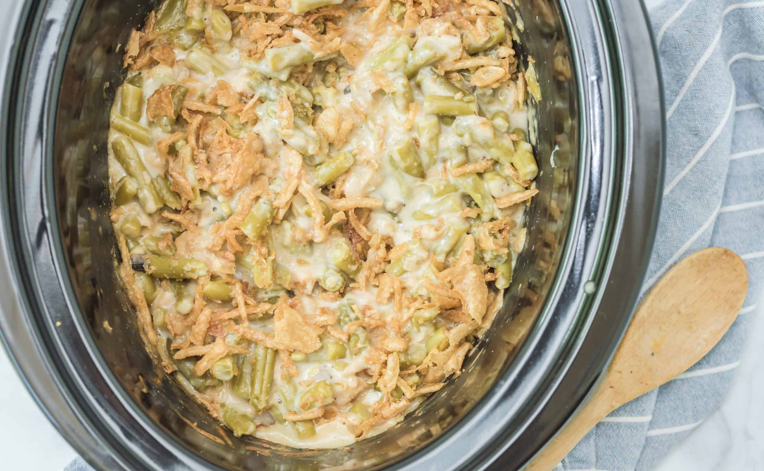 The image features a crockpot containing green bean casserole with a topping of crispy fried onions. Beside the crockpot, there is a wooden spoon and a striped cloth napkin.