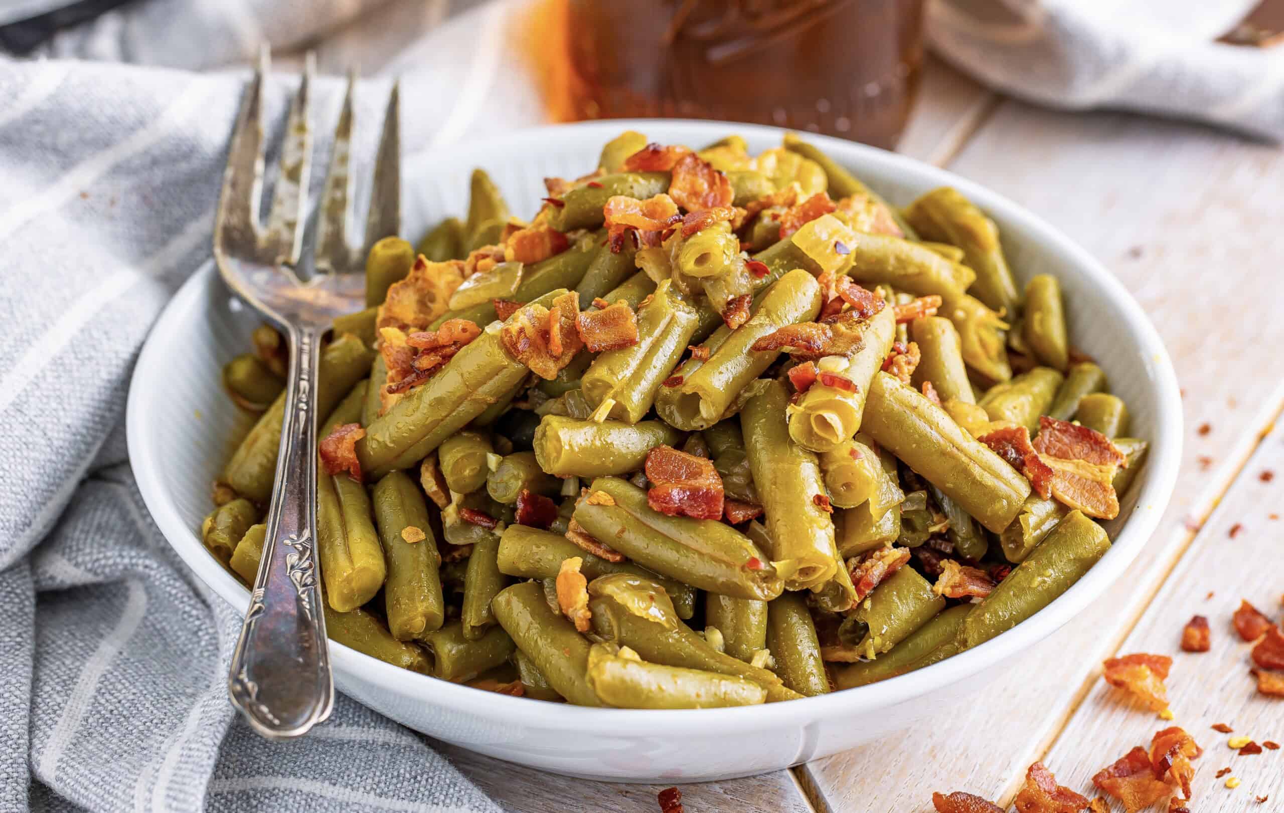 A bowl contains cooked green beans with crispy bacon bits on top, accompanied by a fork resting at its side.
