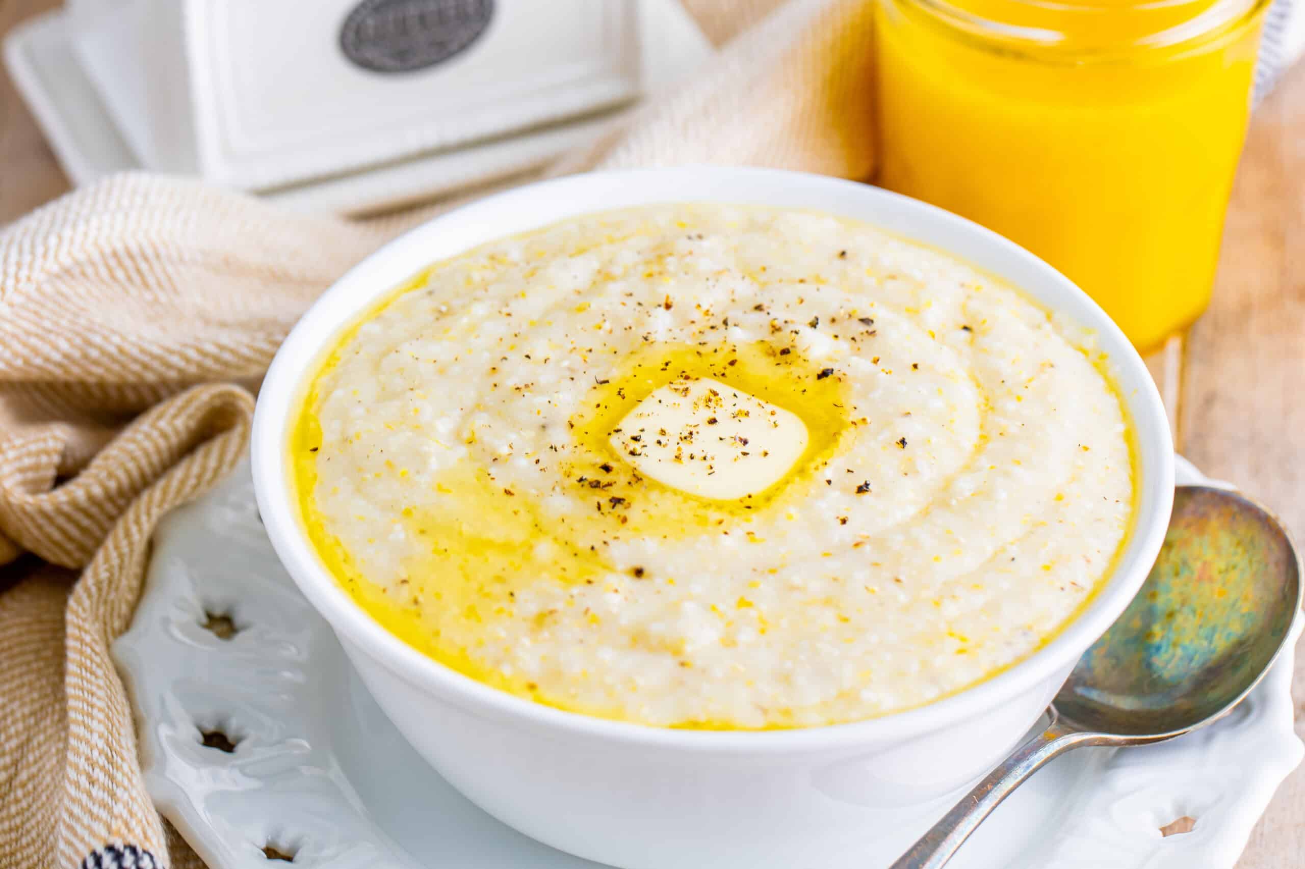 A bowl of creamy grits topped with melted butter and pepper, with a spoon and a jar of an orange-hued beverage in the background.