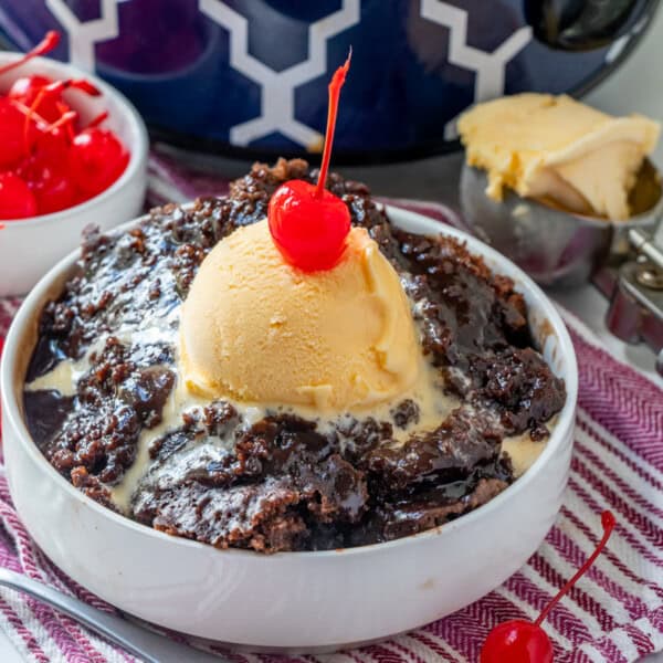 The image includes a bowl of chocolate dessert with a scoop of vanilla ice cream and a cherry on top, a separate bowl filled with cherries, and an ice cream scoop. These items are placed on a red striped cloth.