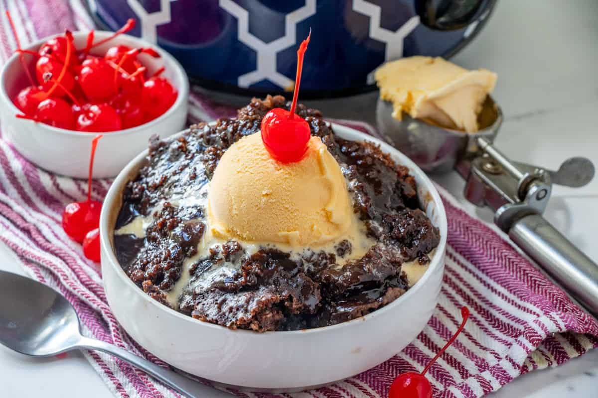 The image includes a bowl of chocolate dessert with a scoop of vanilla ice cream and a cherry on top, a separate bowl filled with cherries, and an ice cream scoop. These items are placed on a red striped cloth.