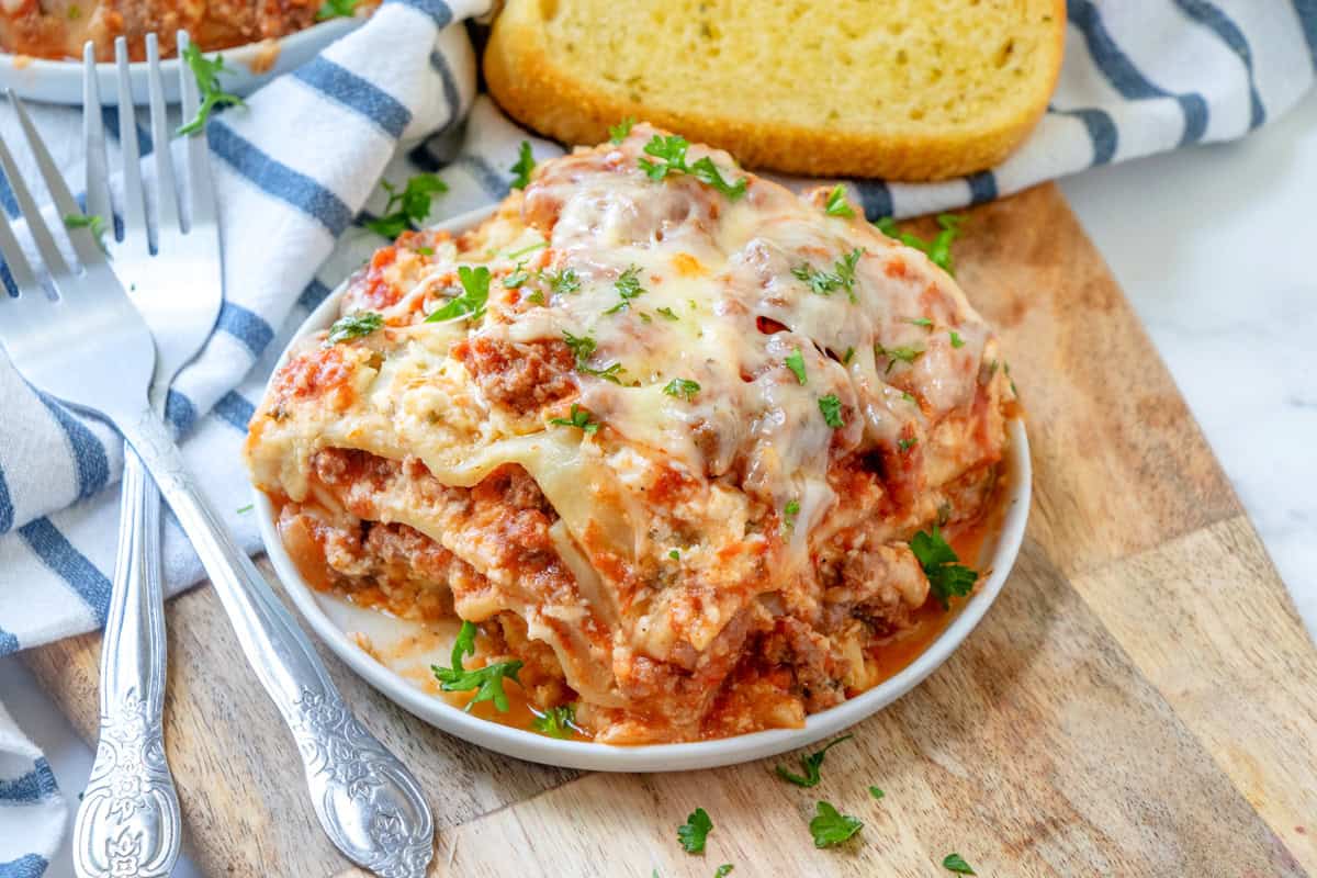 A slice of lasagna topped with melted cheese and parsley sits on a white plate, accompanied by a piece of garlic bread and two forks, all placed on a wooden board.