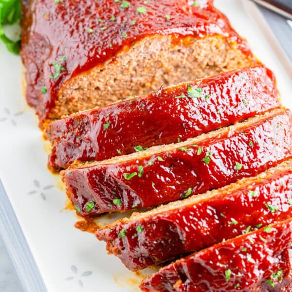 Sliced meatloaf with a glossy glaze on a white platter, garnished with parsley and accompanied by a nearby knife.