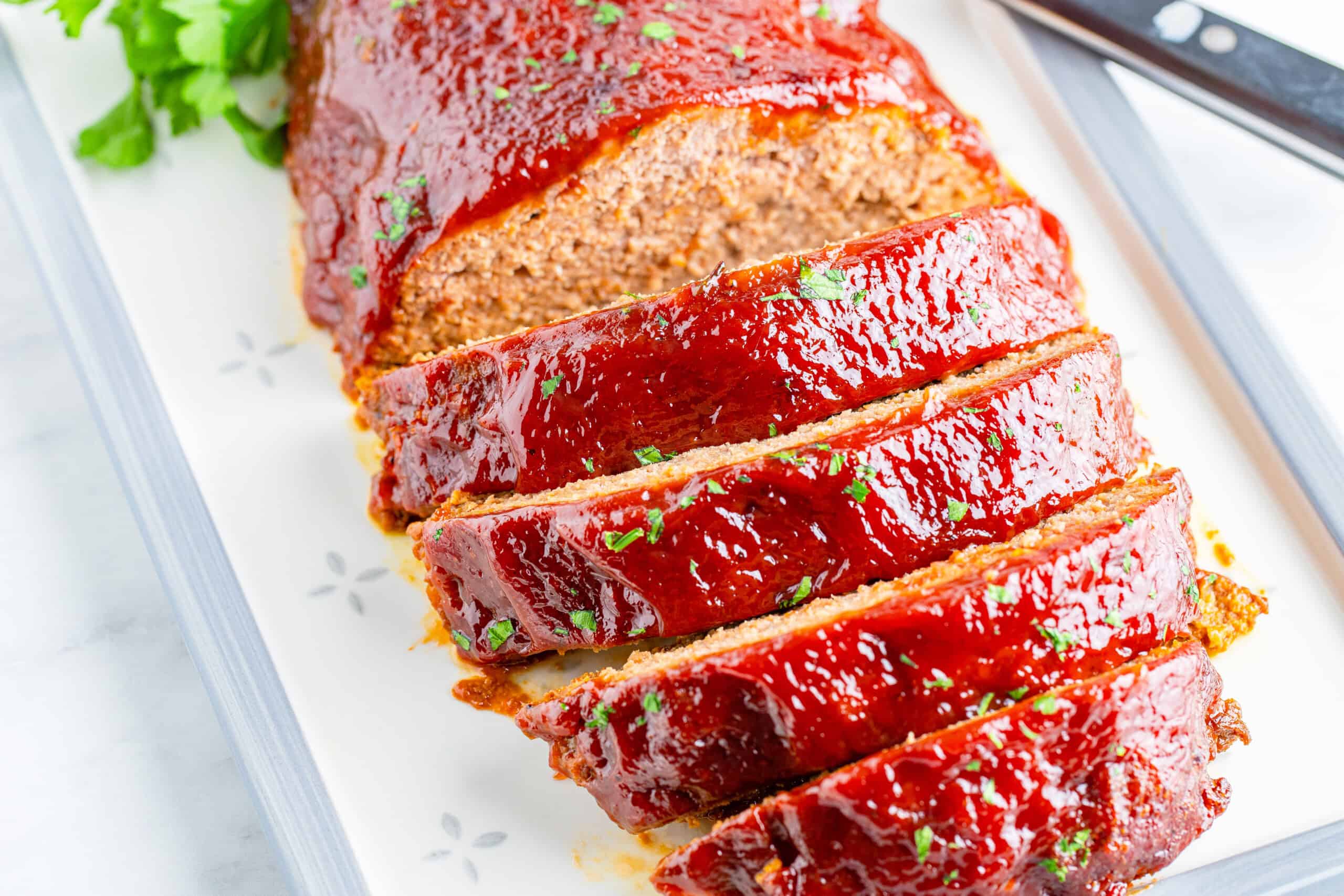 Sliced meatloaf with a glossy glaze on a white platter, garnished with parsley and accompanied by a nearby knife.