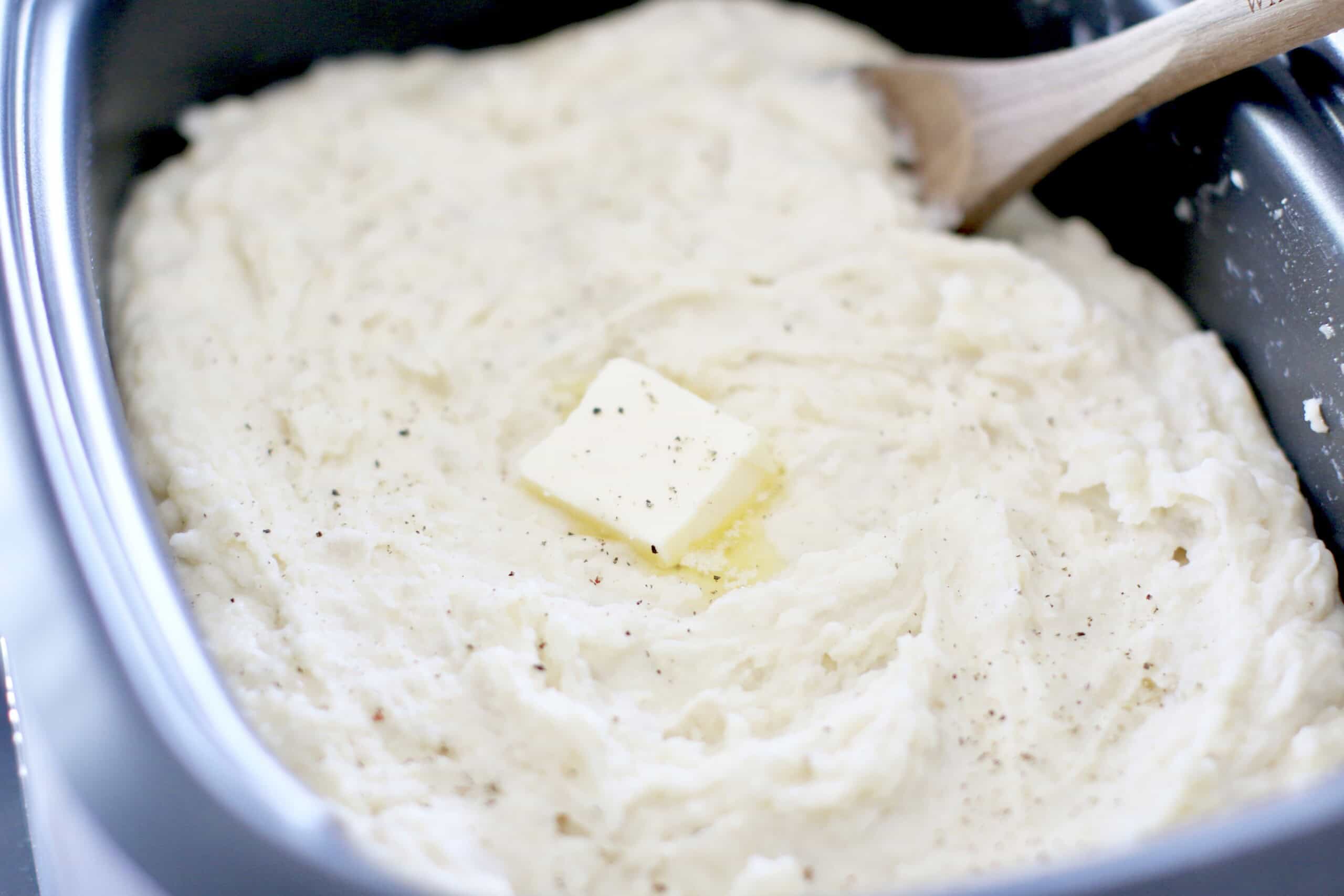 A dish of mashed potatoes with a pat of butter and black pepper, alongside a wooden spoon.