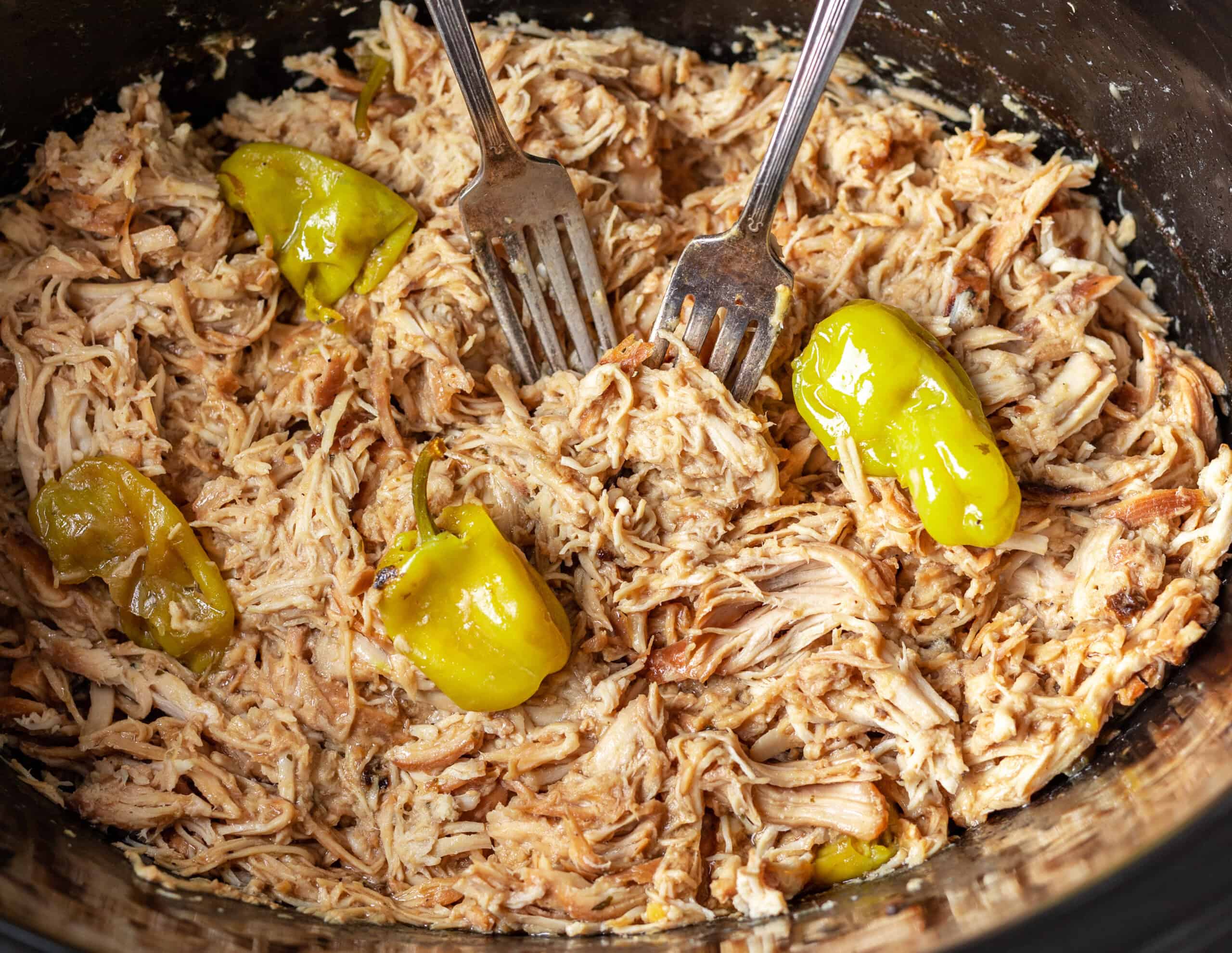 A pot containing shredded chicken and whole pepperoncini peppers, accompanied by two forks.