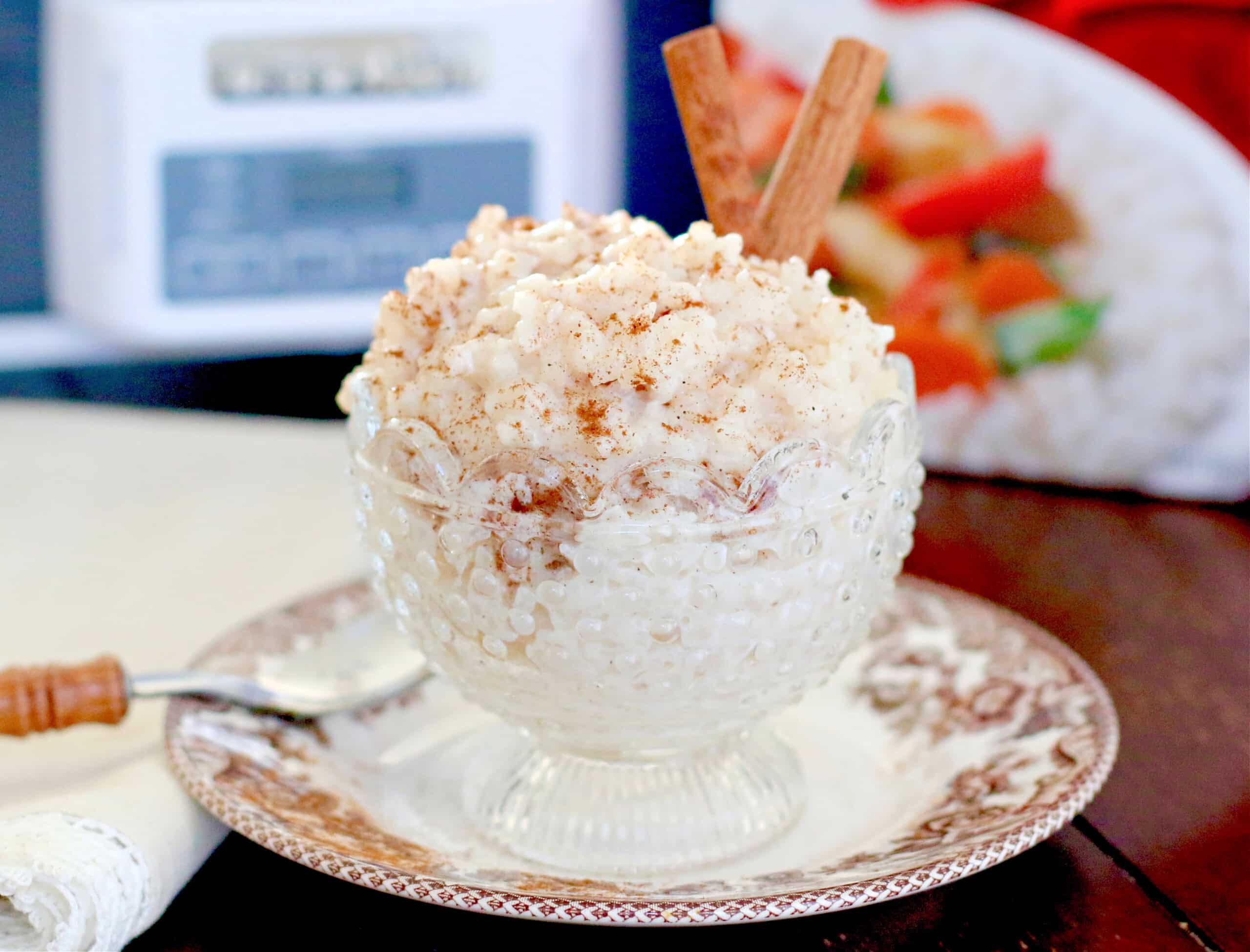 A glass bowl containing rice pudding, garnished with cinnamon and two cinnamon sticks, sits on a decorative saucer with a spoon beside it.