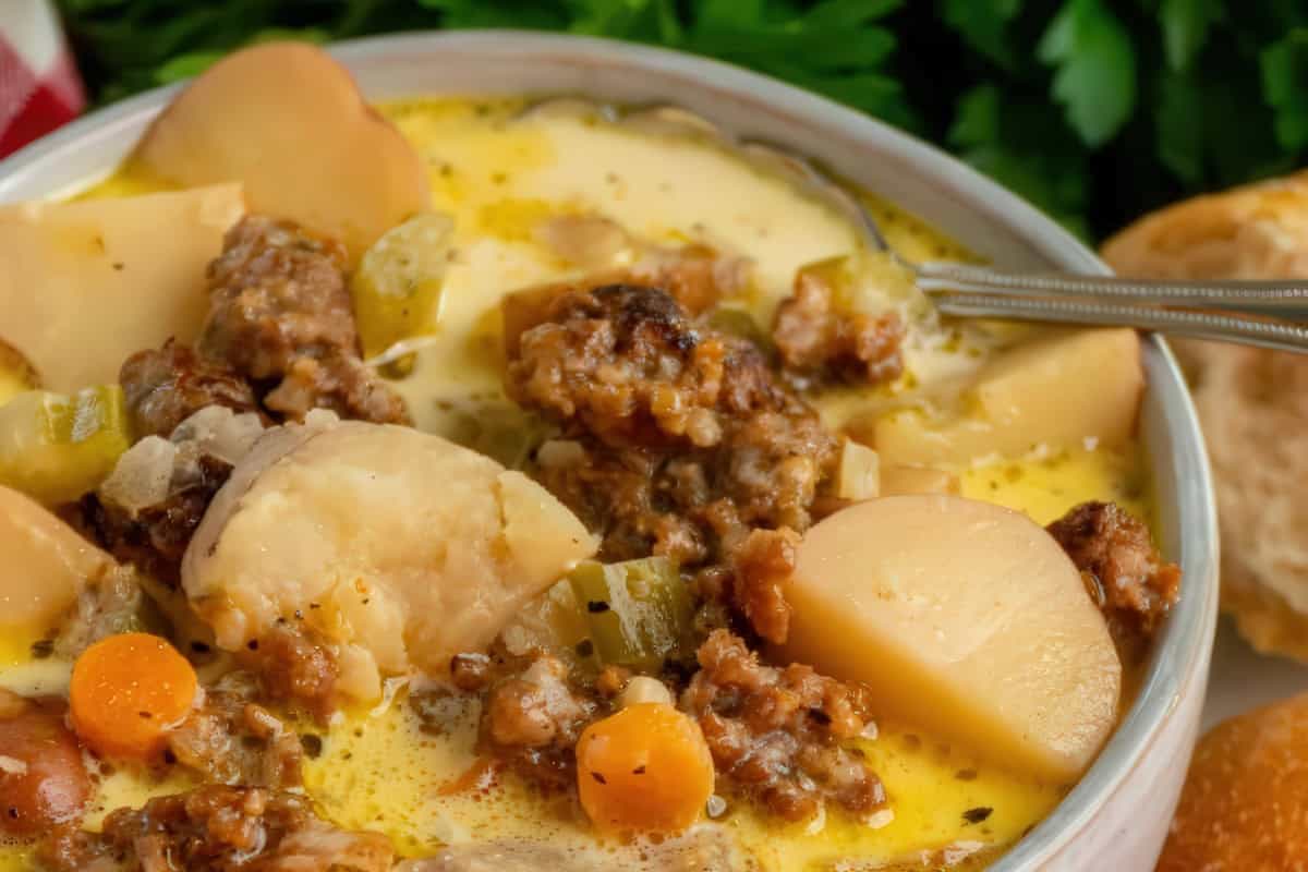 A bowl of creamy soup filled with chunks of potatoes, sausage, carrots, and celery. A spoon is placed on the side of the bowl. Fresh parsley is visible in the background with a slice of bread beside the bowl.