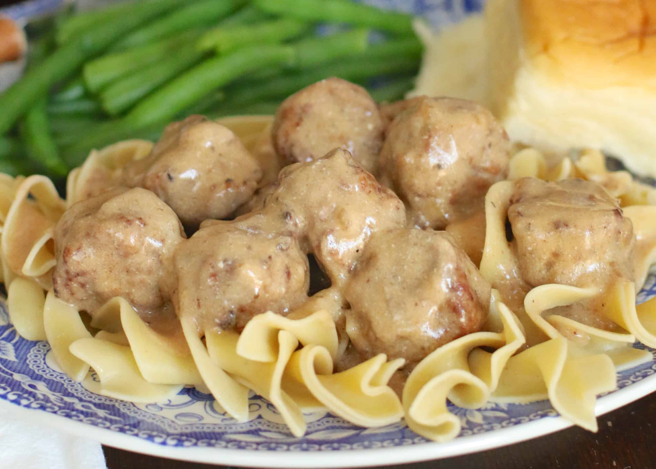 The image depicts Swedish meatballs covered in creamy sauce, served on a bed of egg noodles. Green beans and a roll are also present.