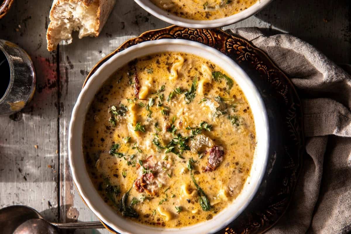 A bowl of creamy soup garnished with herbs and chunks of sausage. The soup is served in a white bowl on a dark, decorative plate, accompanied by a piece of crusty bread. A beige napkin is visible on the side.