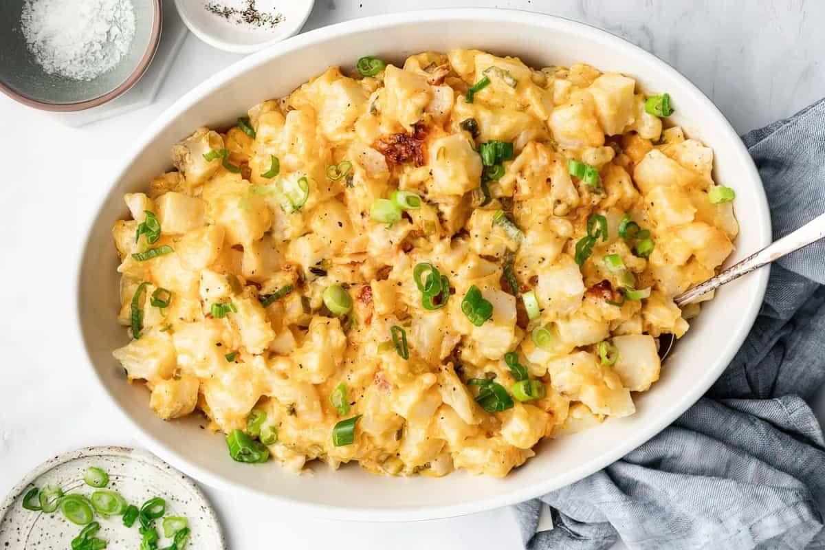 A white oval dish filled with scalloped potatoes topped with chopped green onions, with a spoon inside. Nearby are small bowls containing salt and pepper.