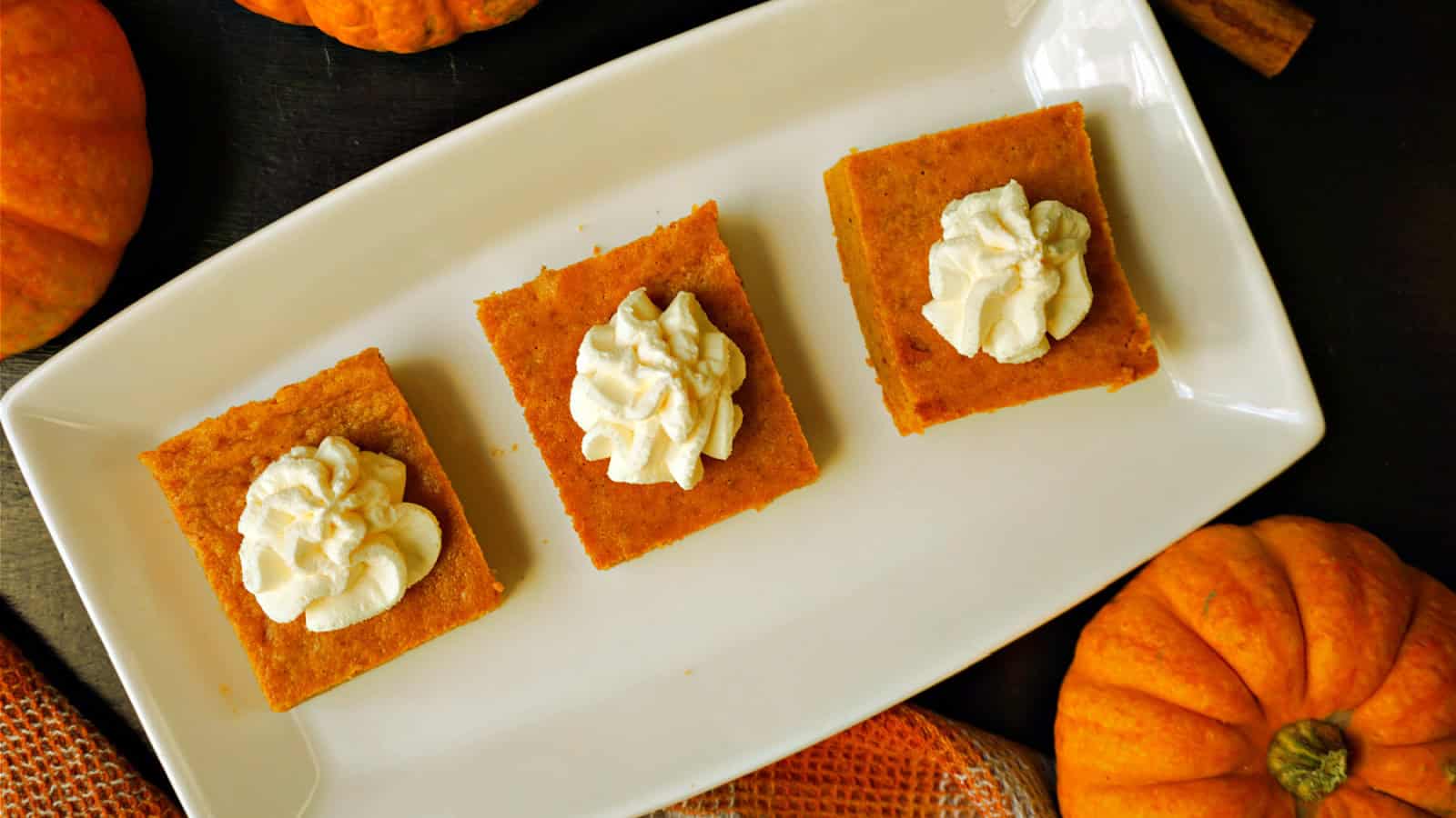 Three pumpkin pie bars topped with whipped cream are arranged on a rectangular white plate. Small pumpkins and an orange napkin are around the plate, set on a dark surface.