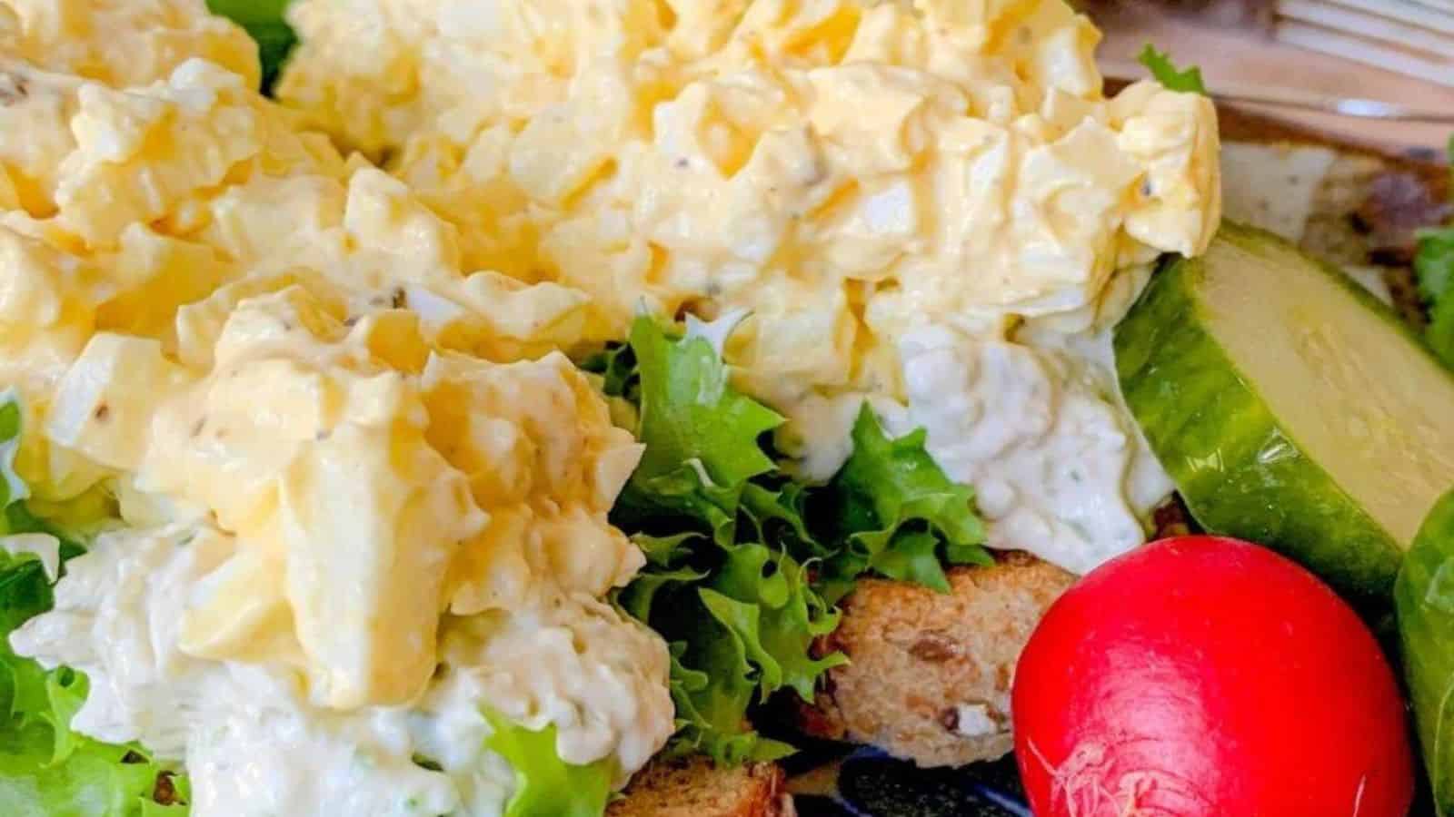 The image features egg salad on pieces of bread, accompanied by leafy greens, sliced cucumbers, and a red radish.