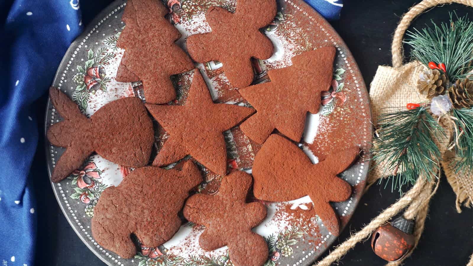 A festive plate filled with various shaped gingerbread cookies, including stars and trees, dusted with powdered sugar. The plate is decorated with a Christmas design, surrounded by a navy cloth with white dots and a pine branch with pinecones.