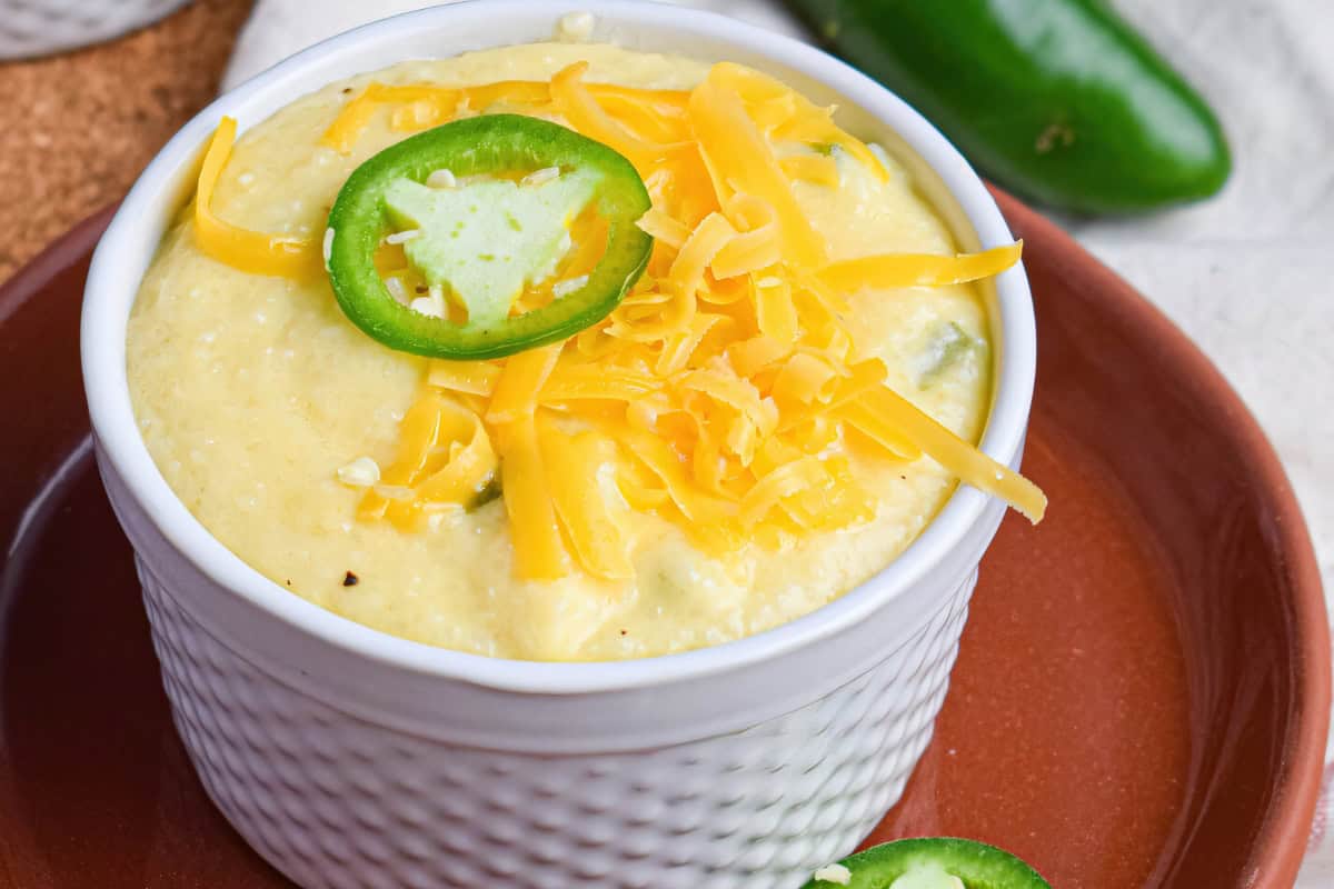 A bowl of creamy grits topped with shredded cheese and sliced jalapeño, placed on a brown plate. In the background, there is a whole jalapeño.