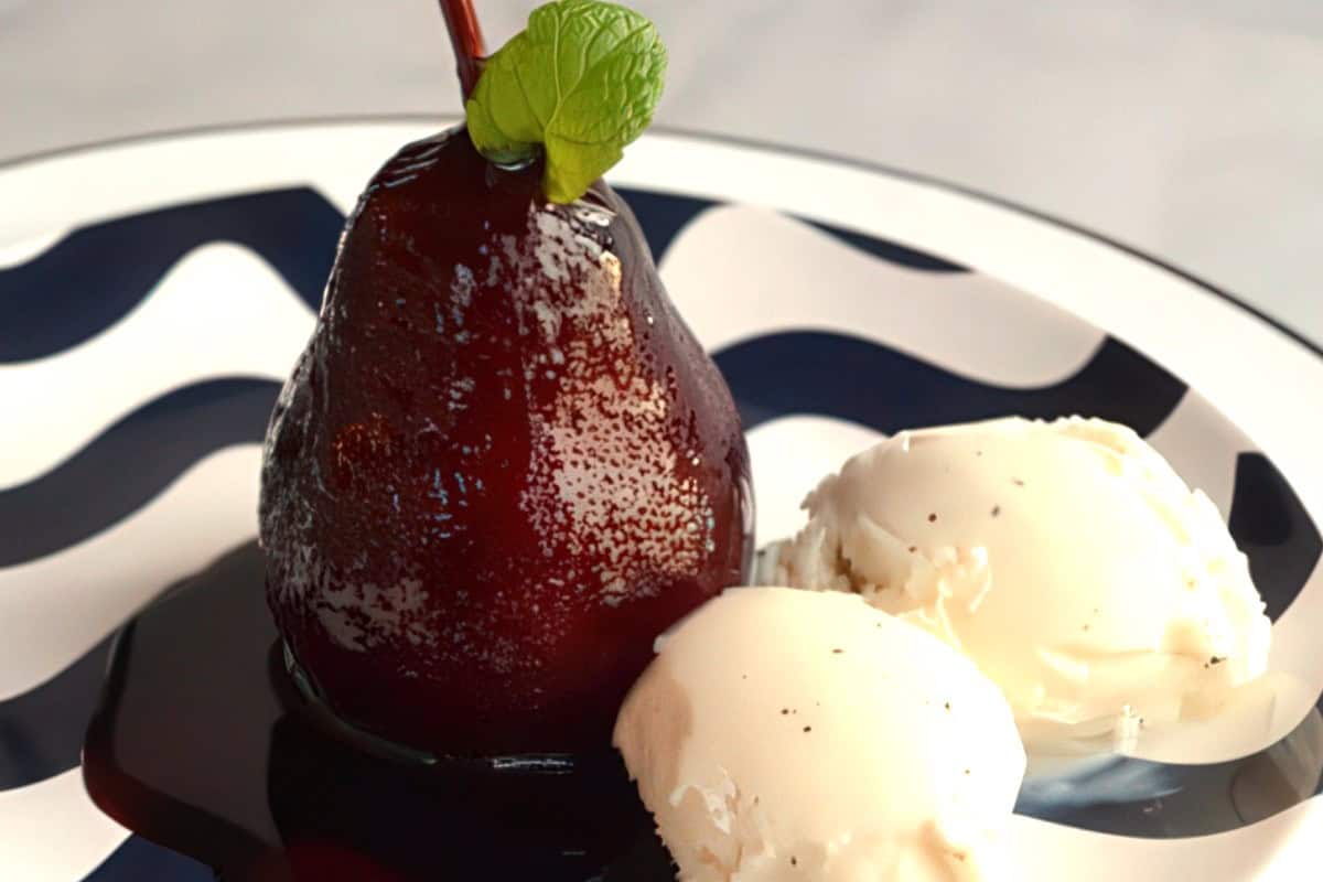A poached pear in red sauce, garnished with a mint leaf, is served on a plate alongside two scoops of vanilla ice cream. The plate has a black and white wavy pattern.