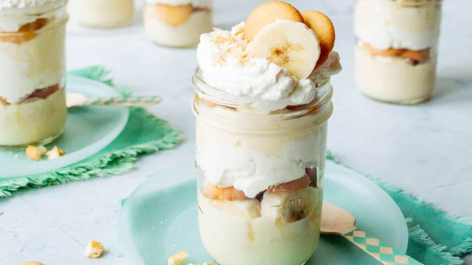 A dessert jar featuring layers of banana pudding, slices of banana, vanilla wafers, and whipped cream, topped with banana slices and wafers. It sits on a light blue plate, with more jars visible in the background.