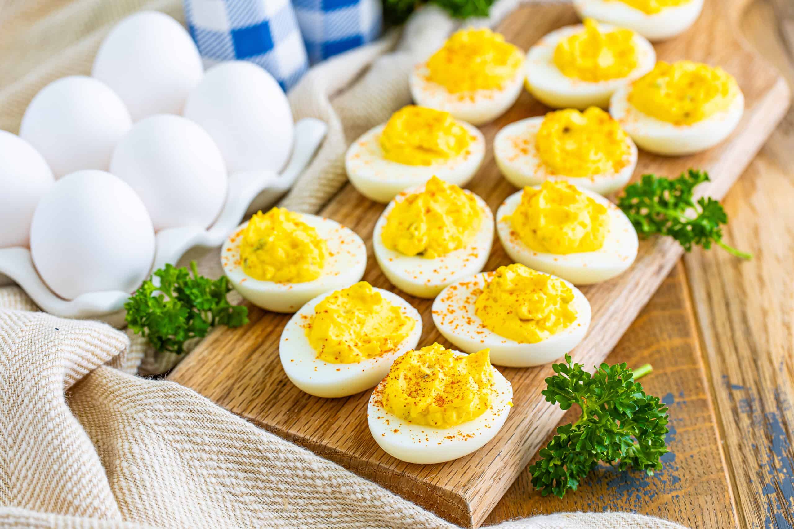 The image features deviled eggs garnished with paprika and parsley on a wooden board, with a carton of whole eggs and a checkered cloth in the background.