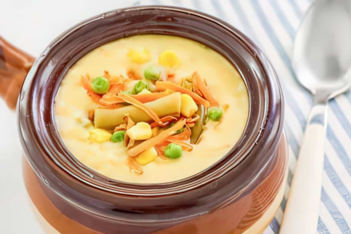 A bowl of creamy soup garnished with corn, peas, and shredded vegetables, placed on a striped cloth next to a spoon.