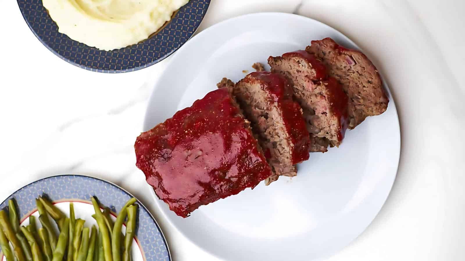 The image shows a sliced meatloaf with a glaze on a white plate, mashed potatoes in one dish, and green beans in another.