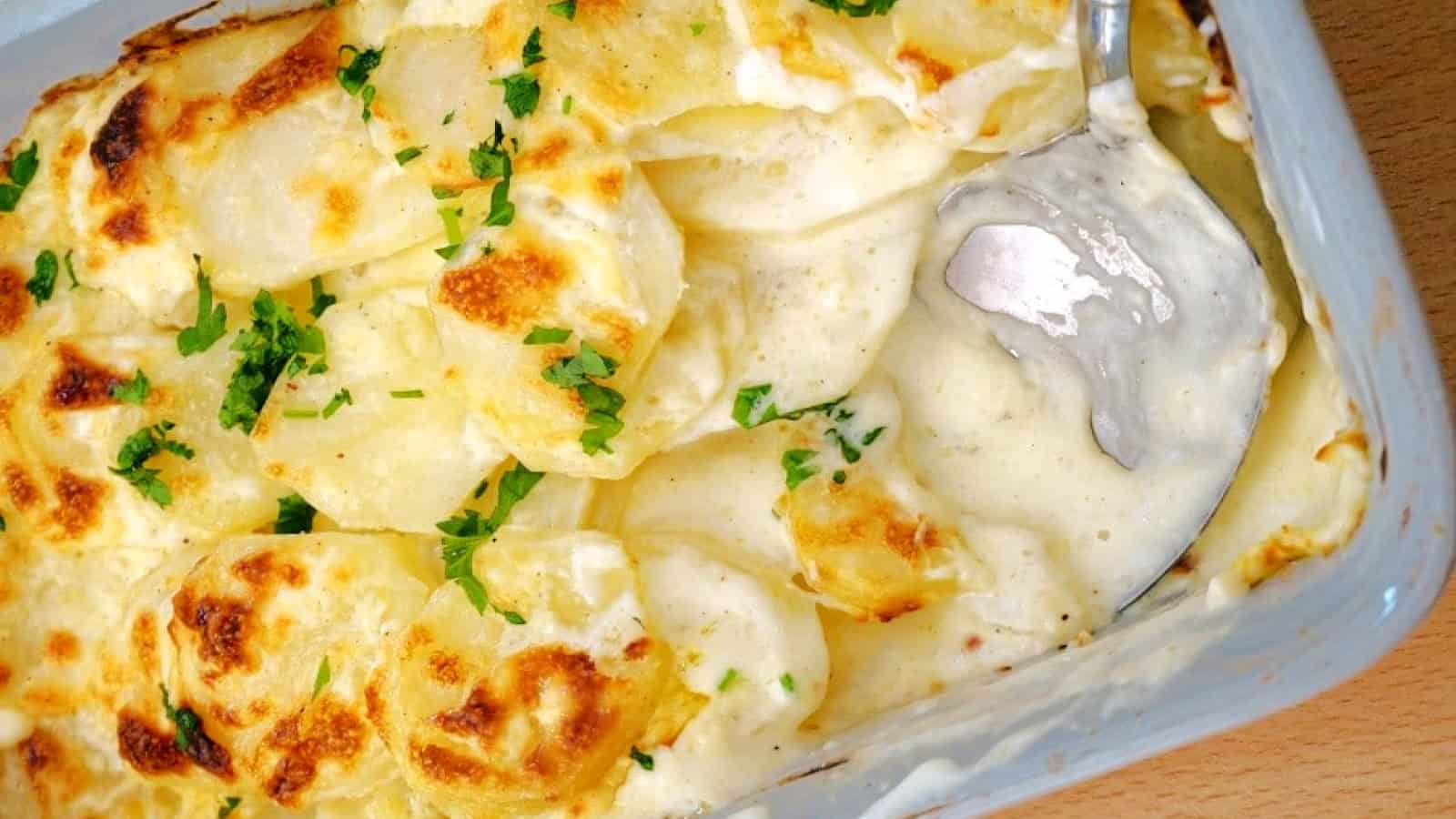 A close-up image of a dish filled with creamy scalloped potatoes, topped with sprigs of parsley and accompanied by a serving spoon inserted on the side.