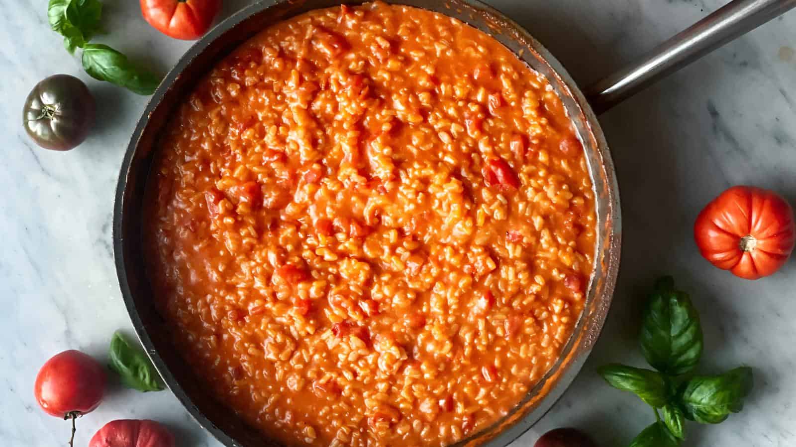 A pan filled with creamy tomato risotto, surrounded by fresh tomatoes and basil leaves on a marble surface.
