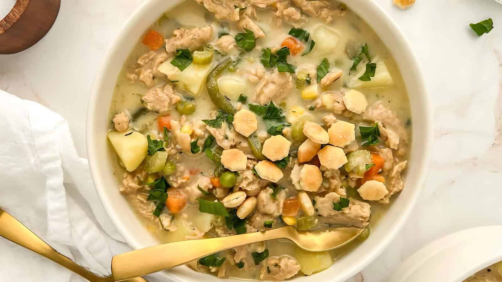 A bowl of creamy chicken soup with vegetables and parsley, topped with round crackers, placed on a white marble surface alongside a golden spoon.