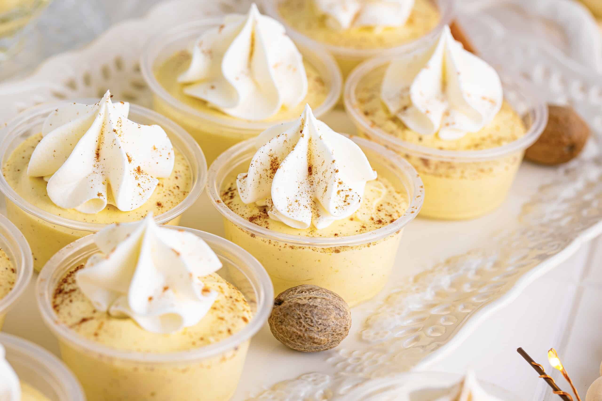The image includes small dessert cups topped with whipped cream and a sprinkle of spice, arranged on a white tray. Whole nutmeg is placed alongside the cups.