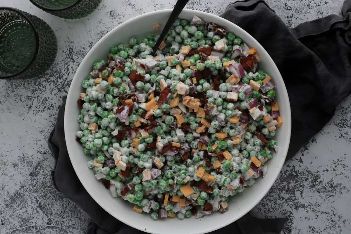 A bowl filled with diced ham, cheese, red onions, and peas in a creamy dressing, set on a textured surface with green glassware adjacent.