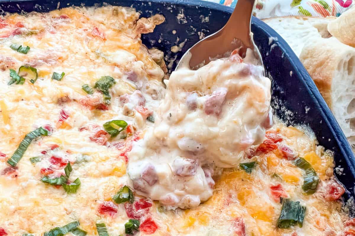 A close-up of a creamy, melted cheese dish with chopped tomatoes and green onions in a dark baking dish. A spoon is scooping a portion, revealing a thick, gooey texture. Bread is visible in the background.
