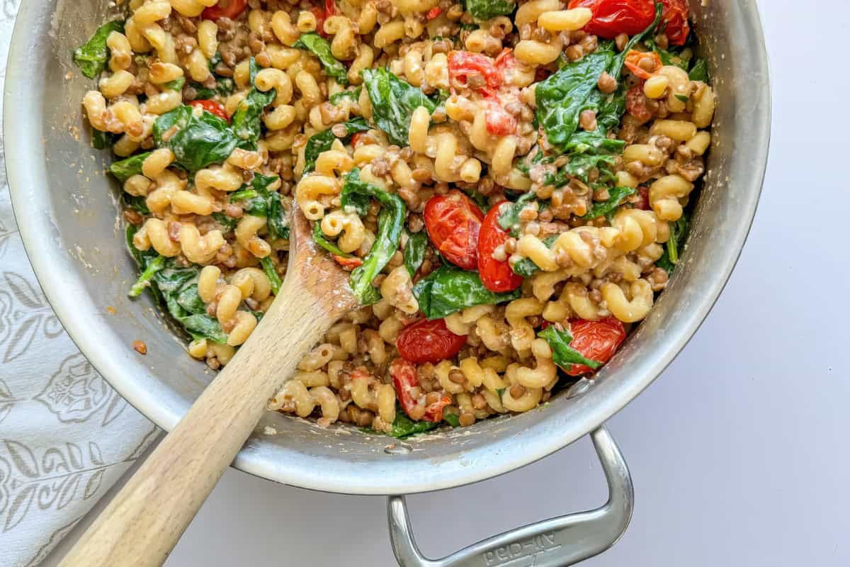 A pan filled with spiral noodles, cherry tomatoes, spinach, and lentils, with a wooden spoon inside.