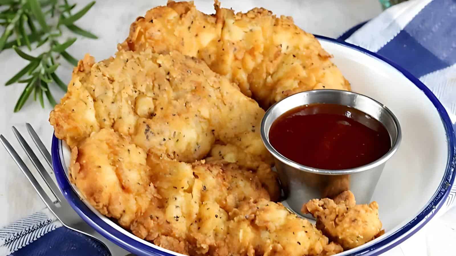 Close-up of a plate with crispy, golden-brown chicken tenders next to a small metal cup of barbecue sauce. A fork and a blue-and-white striped napkin are partially visible on the side.